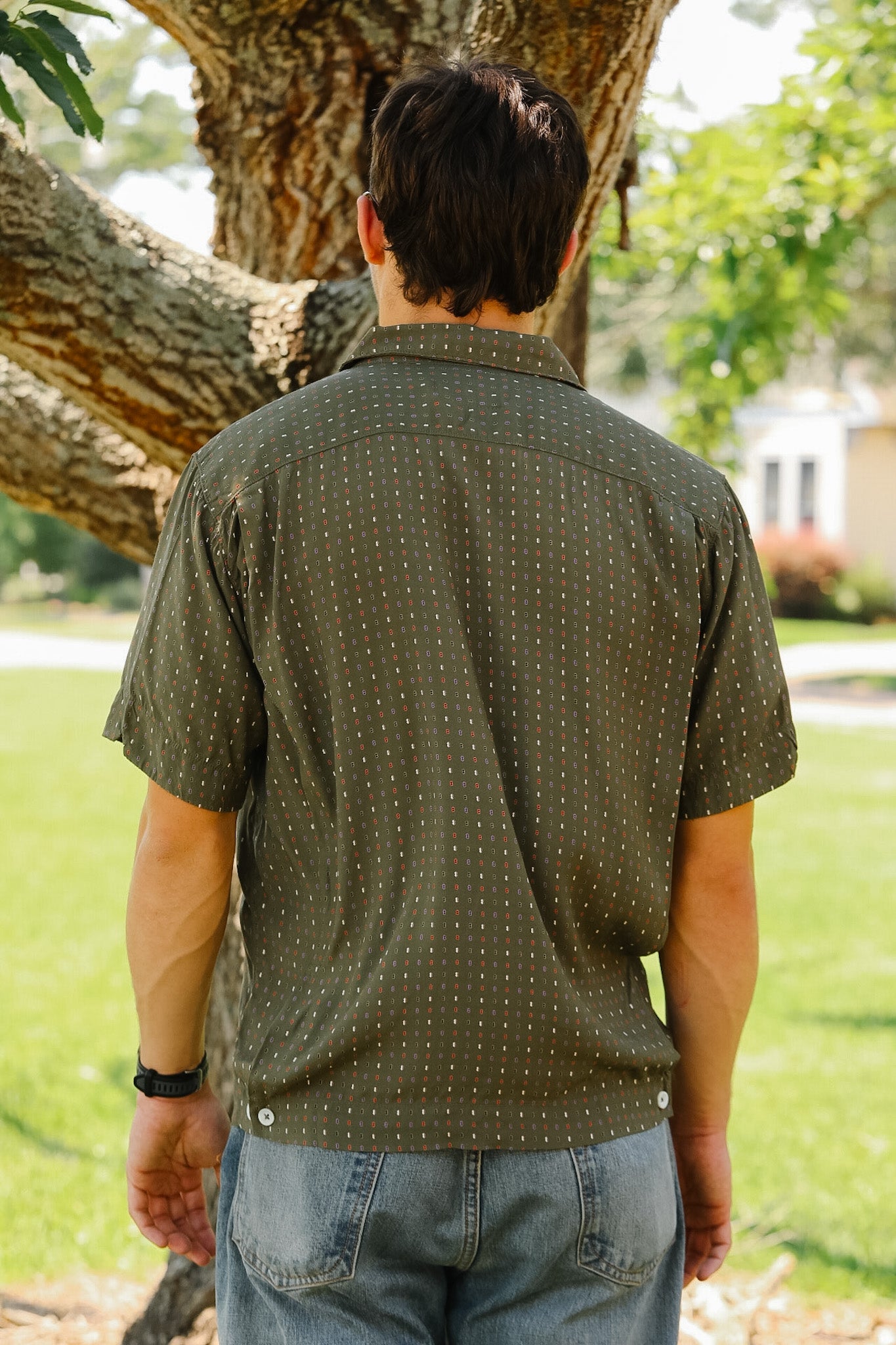 Man wearing a green patterned shirt and blue jeans standing outdoors near a tree