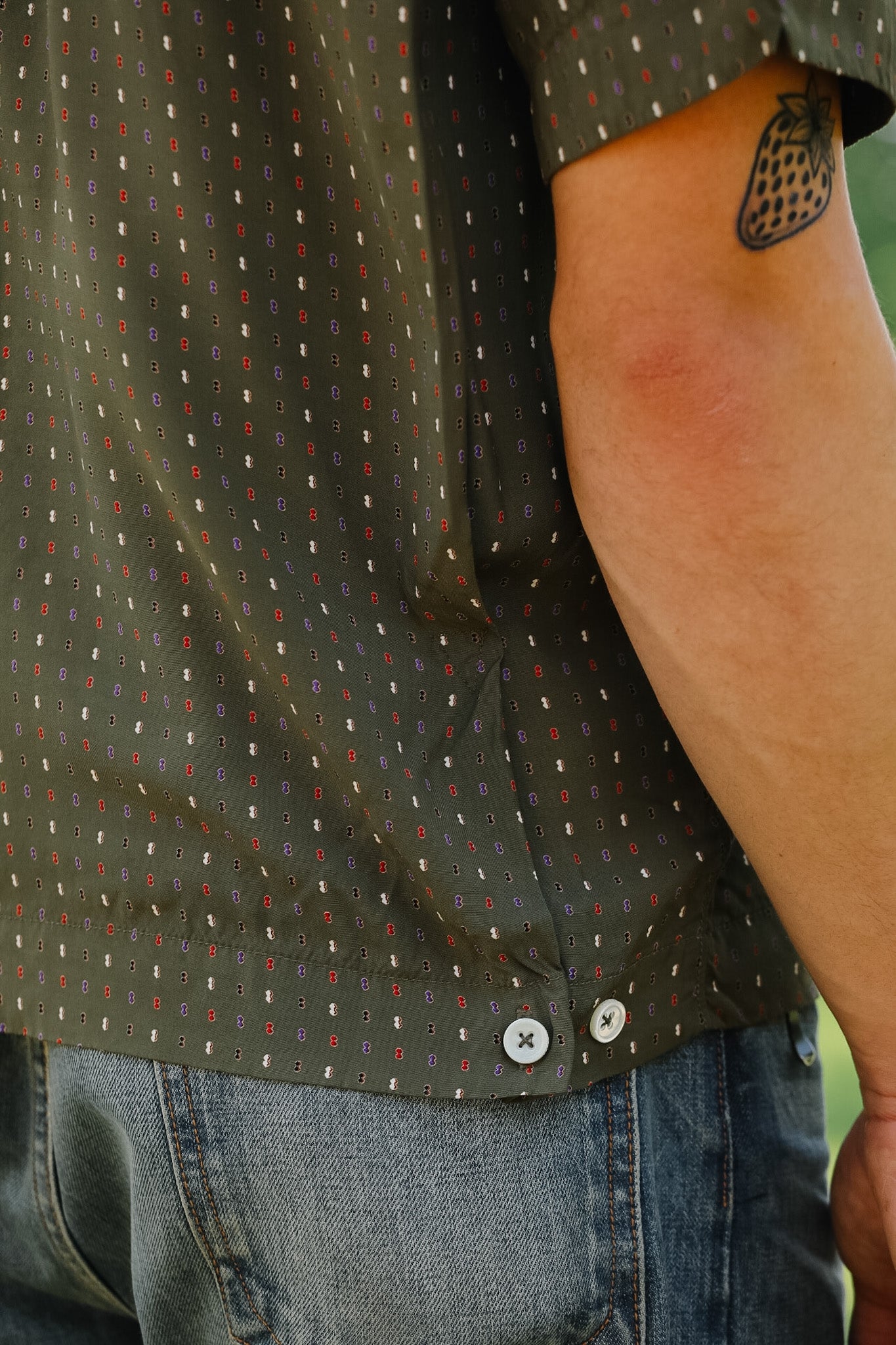 Person wearing a green patterned shirt and blue jeans with a tattoo on their arm.