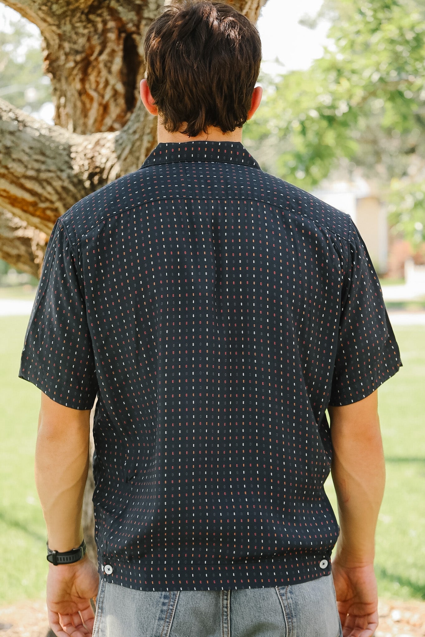Person wearing a dark patterned shirt standing outdoors with trees in the background