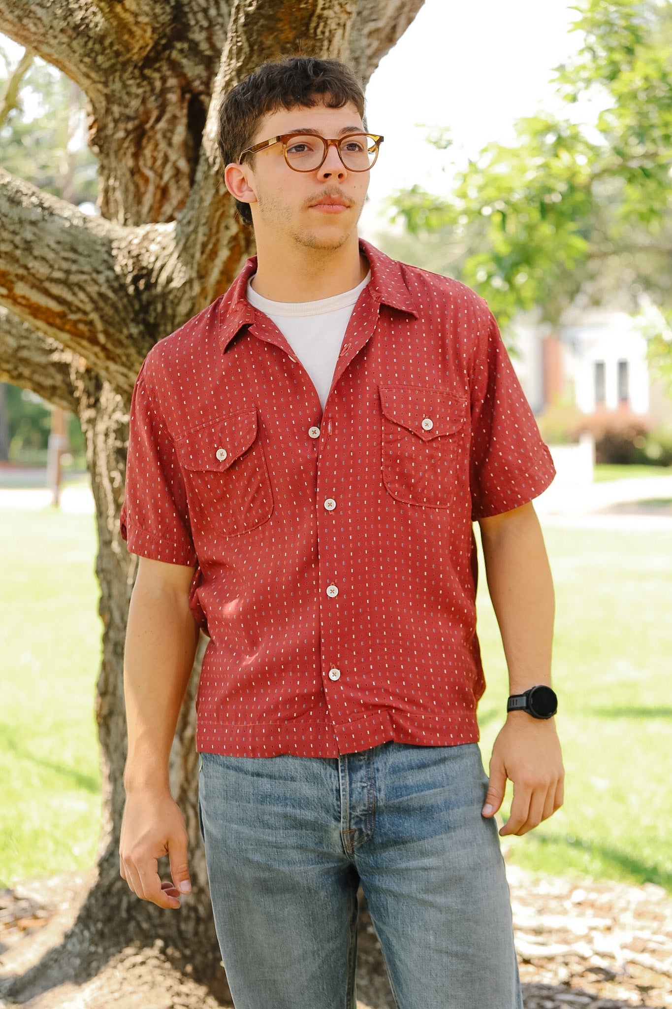 Man wearing a red patterned shirt and blue jeans standing under a tree.