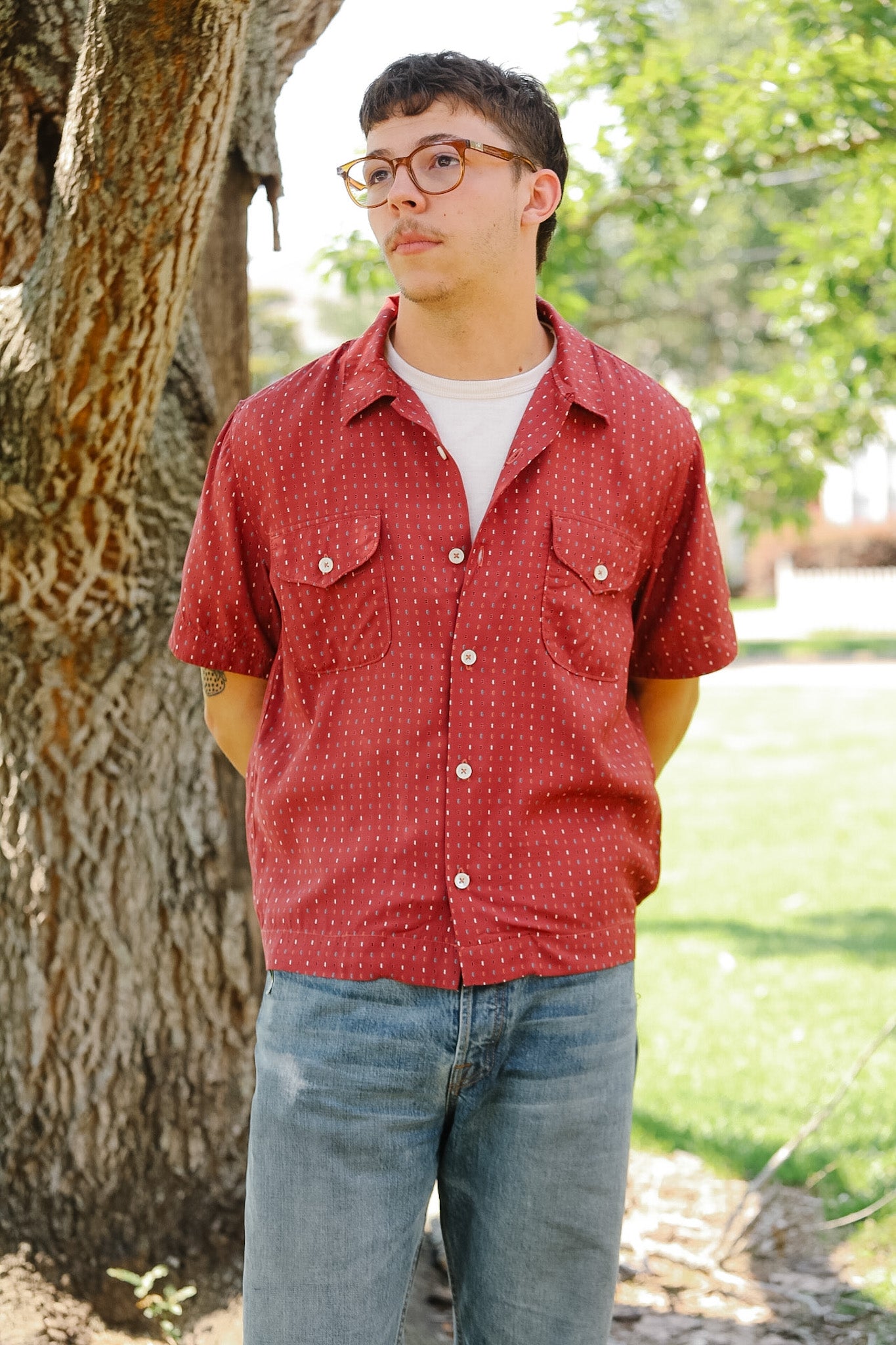 Person wearing a red patterned shirt and blue jeans standing outdoors near a tree.