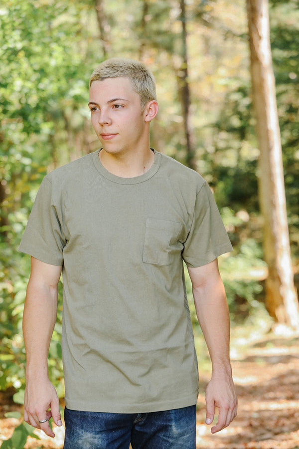 Young man outdoors wearing olive green pocket t-shirt and blue jeans in forest setting