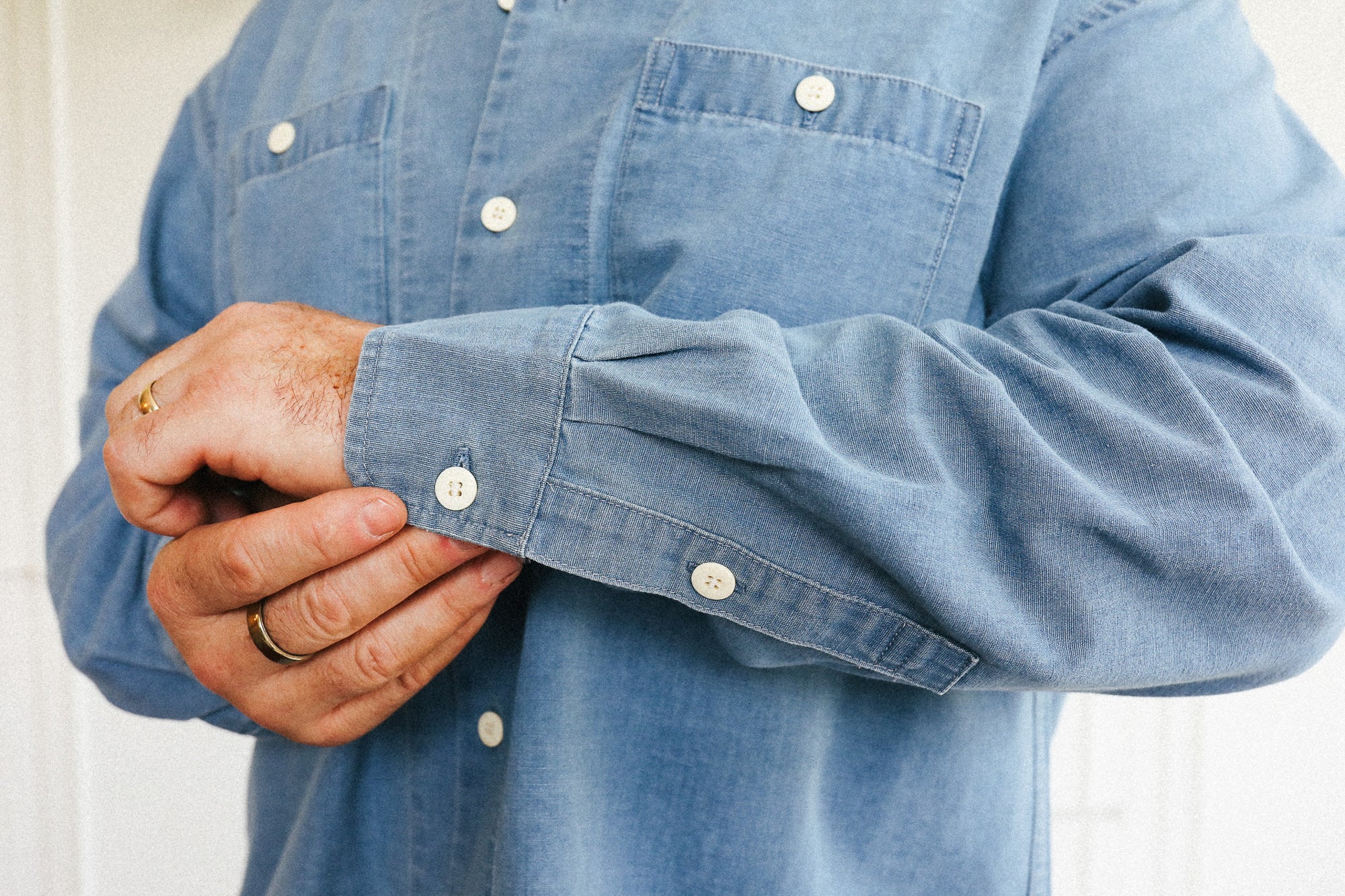 A close-up of a person wearing a light blue chambray button-down shirt, adjusting the cuff with both hands. The shirt features a classic collarless pocket design and white buttons.