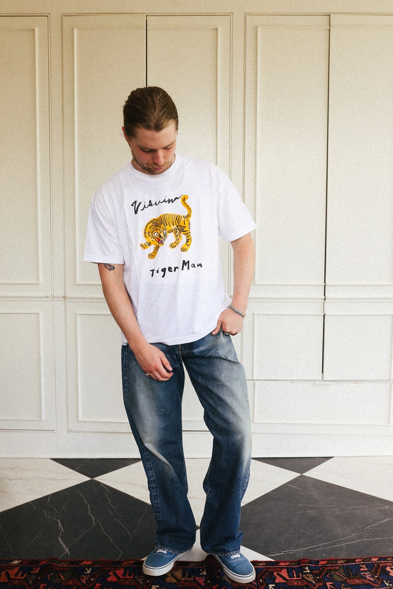 A man wearing a white T-shirt with a yellow and black tiger graphic and the text 'William Tiger Man', paired with blue jeans and blue sneakers, standing in front of a white paneled wall.