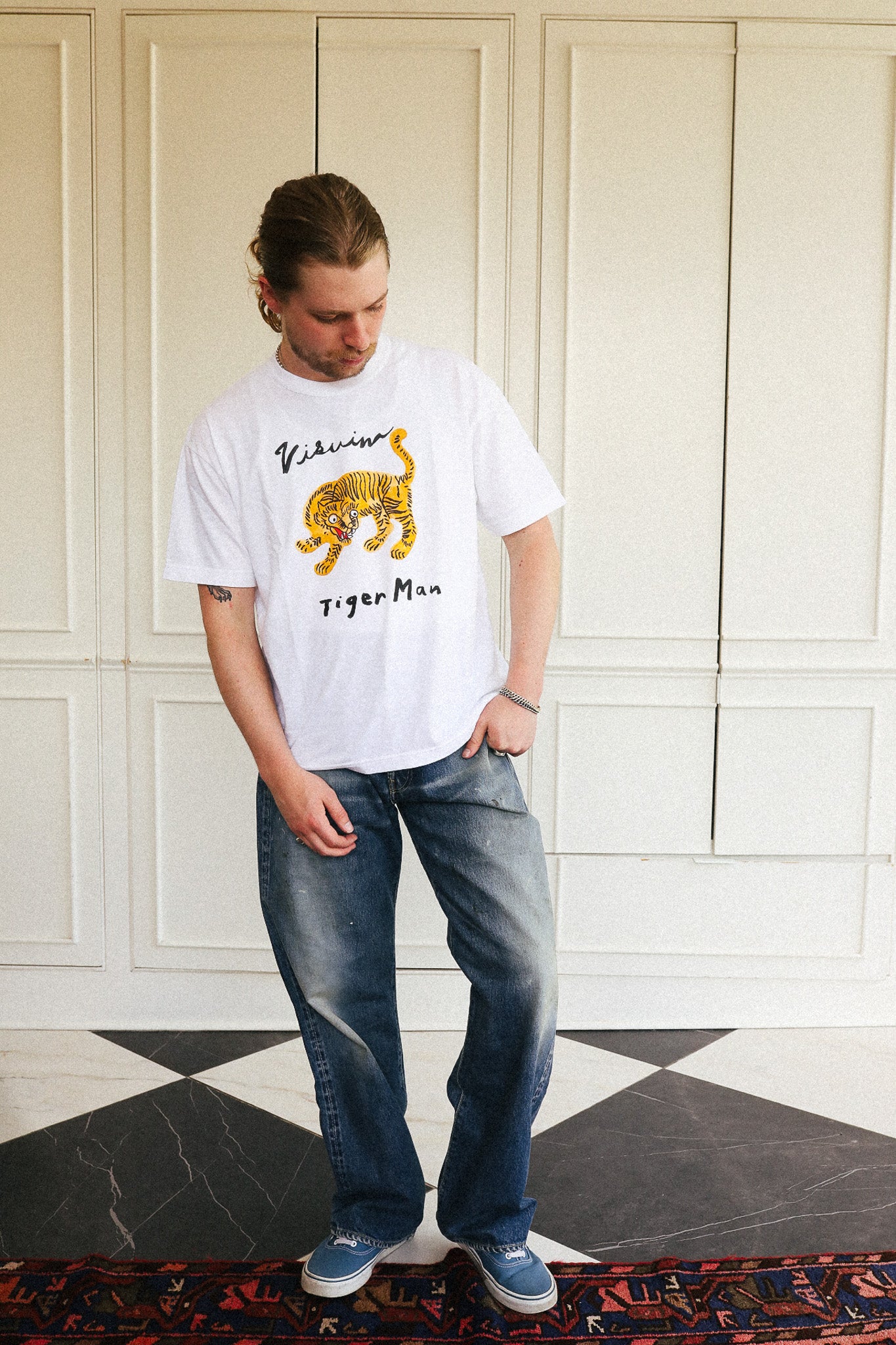 A man wearing a white T-shirt with a yellow and black tiger graphic and the text 'Vilaine Tiger Man', paired with blue jeans and blue sneakers, standing in front of a cream-colored paneled wall.