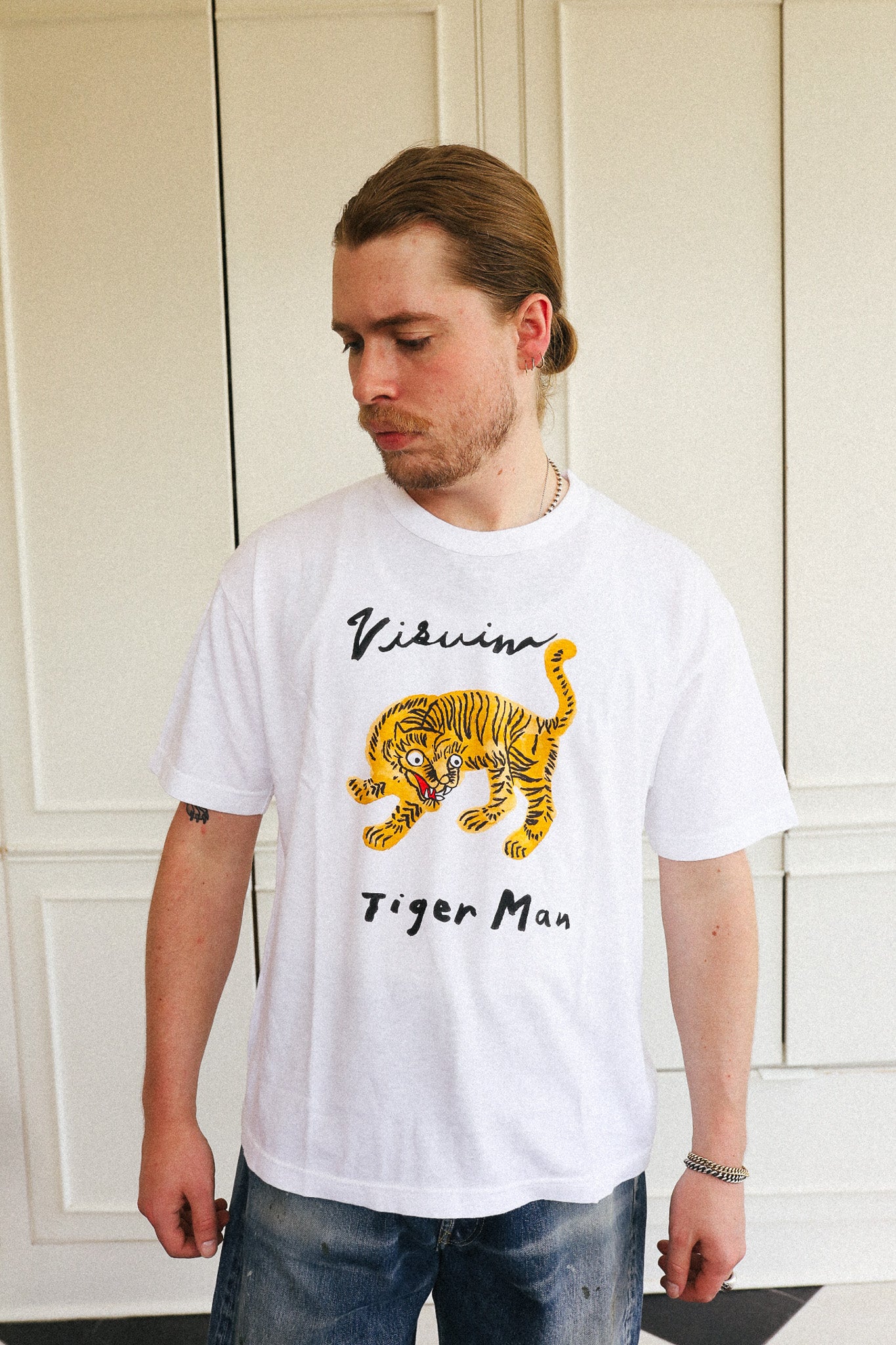 A man wearing a white short-sleeve t-shirt with a yellow and black tiger graphic and the text 'Visvim Tiger Man' printed on the front, paired with blue jeans, standing in front of a cream-colored paneled door.