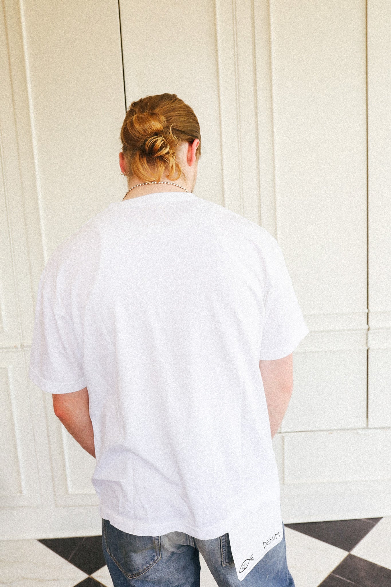 A person viewed from behind wearing a white oversized short-sleeve t-shirt and blue jeans, standing in front of a cream paneled wall.