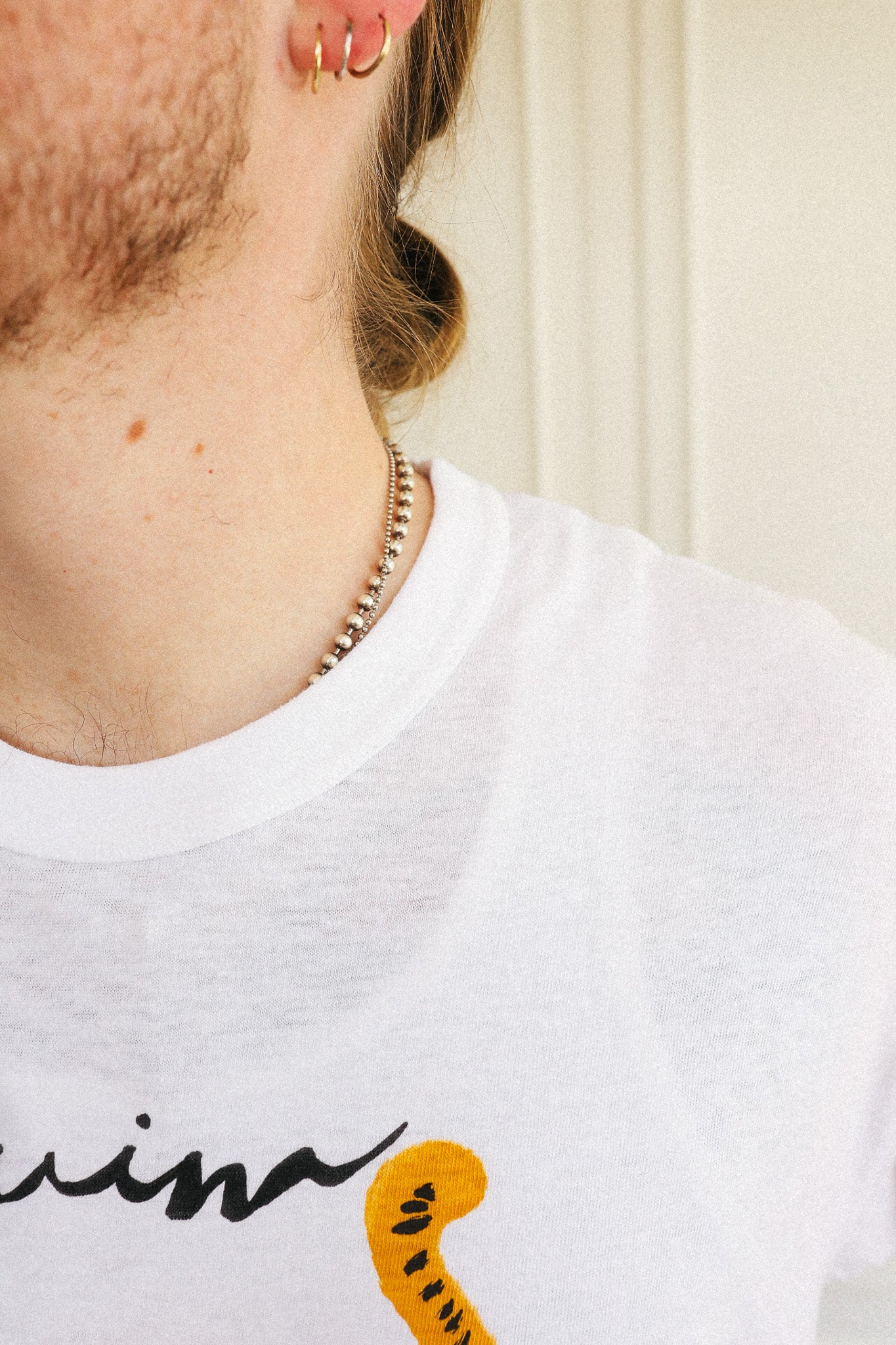 Close-up of a person wearing a white crew-neck t-shirt with a graphic print, accessorized with a gold chain necklace and hoop earrings.