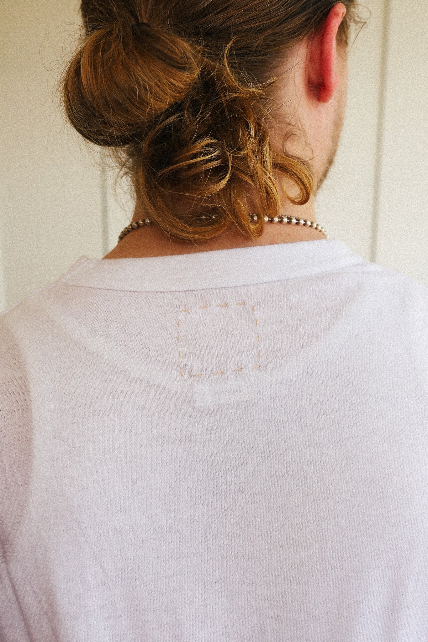 Back view of a person wearing a light gray heather crewneck t-shirt, showing a square patch of unfinished embroidery on the upper back.