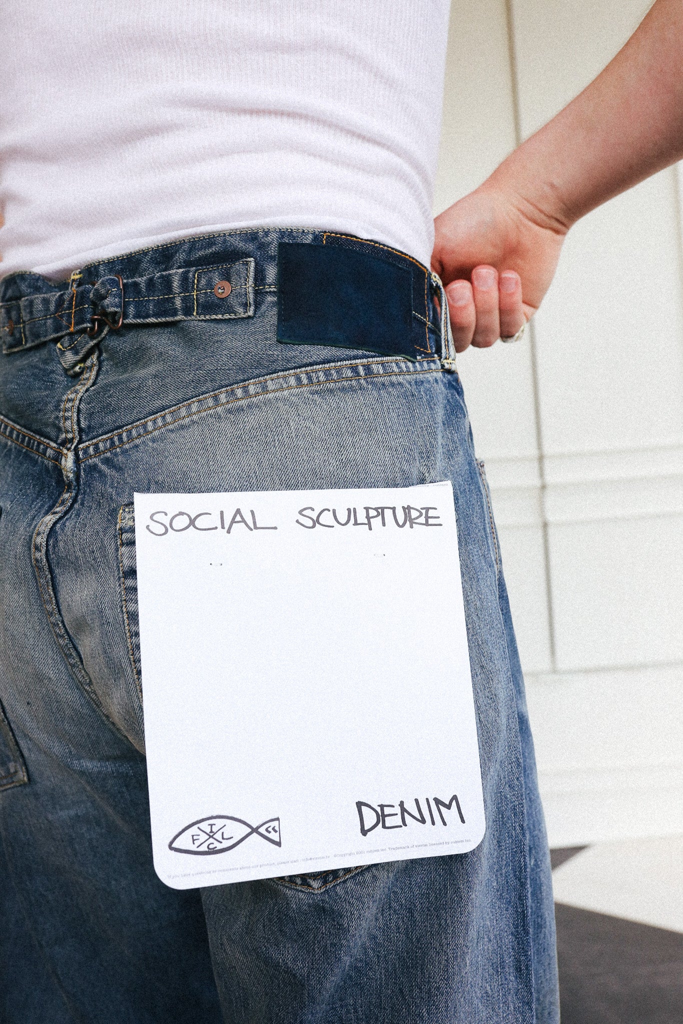 A close-up of a person wearing blue denim jeans with a white rectangular note pinned or attached to the back pocket. The note reads 'SOCIAL SCULPTURE' at the top and 'DENIM' at the bottom, with a small fish-like logo.
