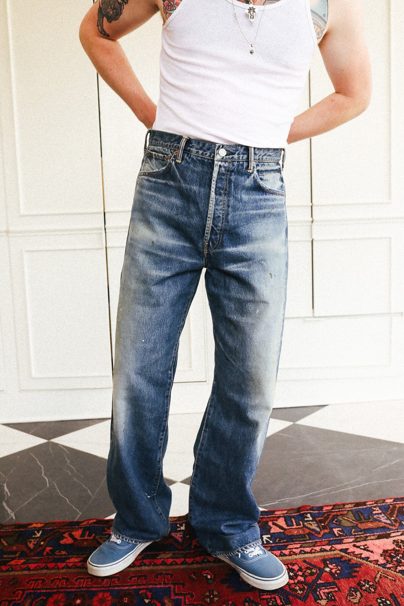 A person wearing a pair of faded blue wide-leg jeans, a white tank top, and blue sneakers, standing indoors on a patterned rug in front of a white paneled wall.