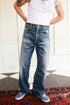 A person wearing a pair of faded blue wide-leg jeans, a white tank top, and blue sneakers, standing indoors on a patterned rug in front of a white paneled wall.