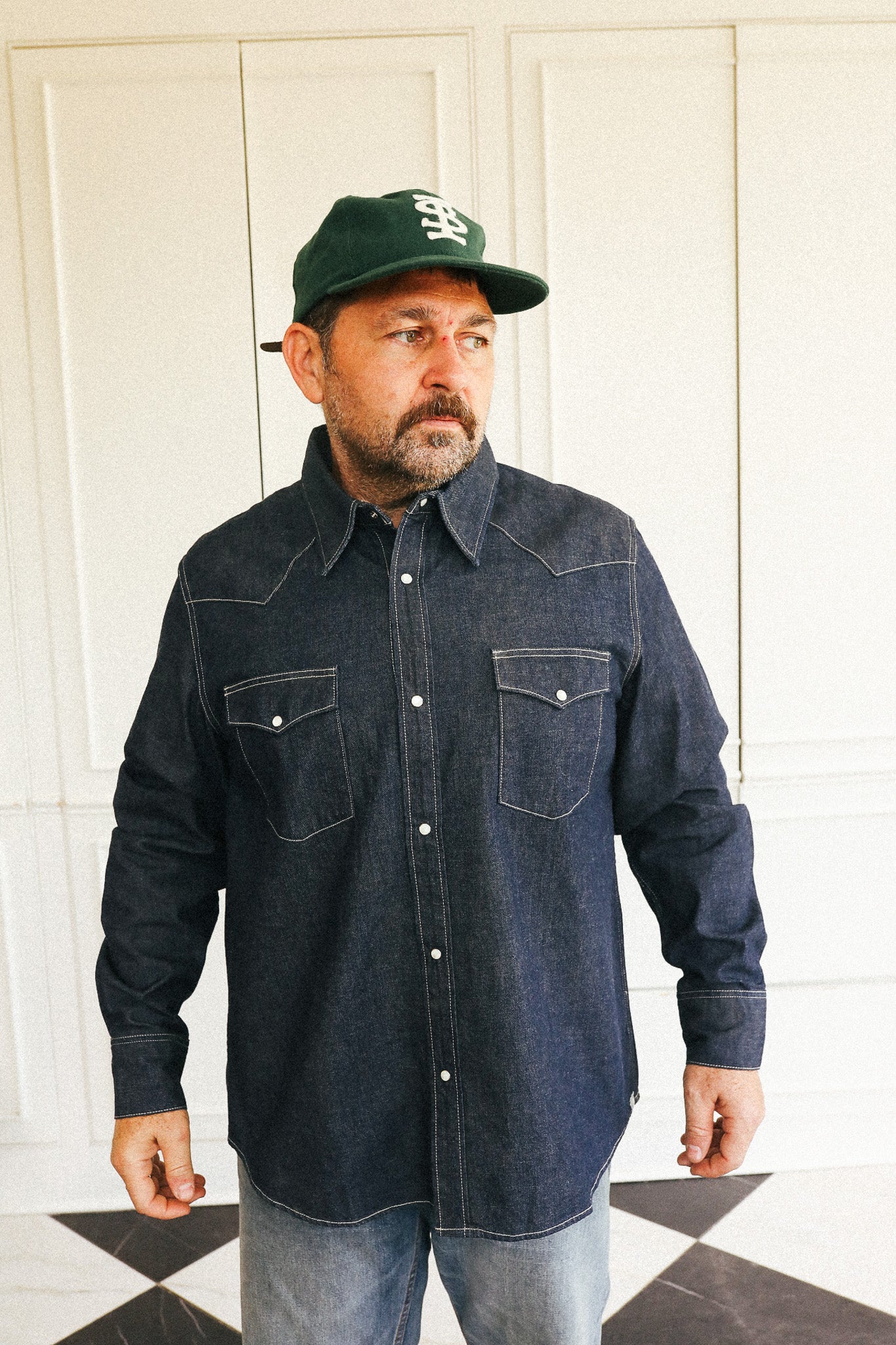 A man wearing a dark indigo denim western-style button-down shirt with contrast stitching, paired with a green baseball cap and light-wash jeans, standing in front of a white paneled wall.