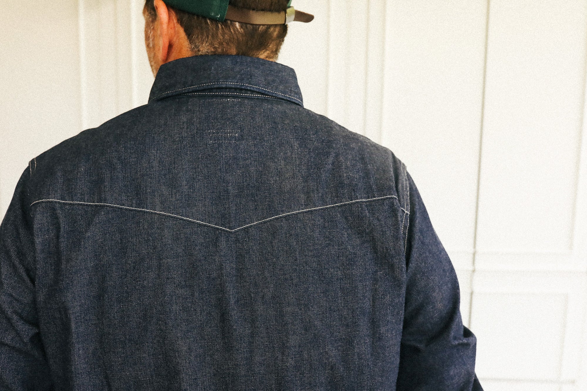 Back view of a man wearing a dark indigo denim shirt jacket with contrast stitching, standing against a white paneled door.