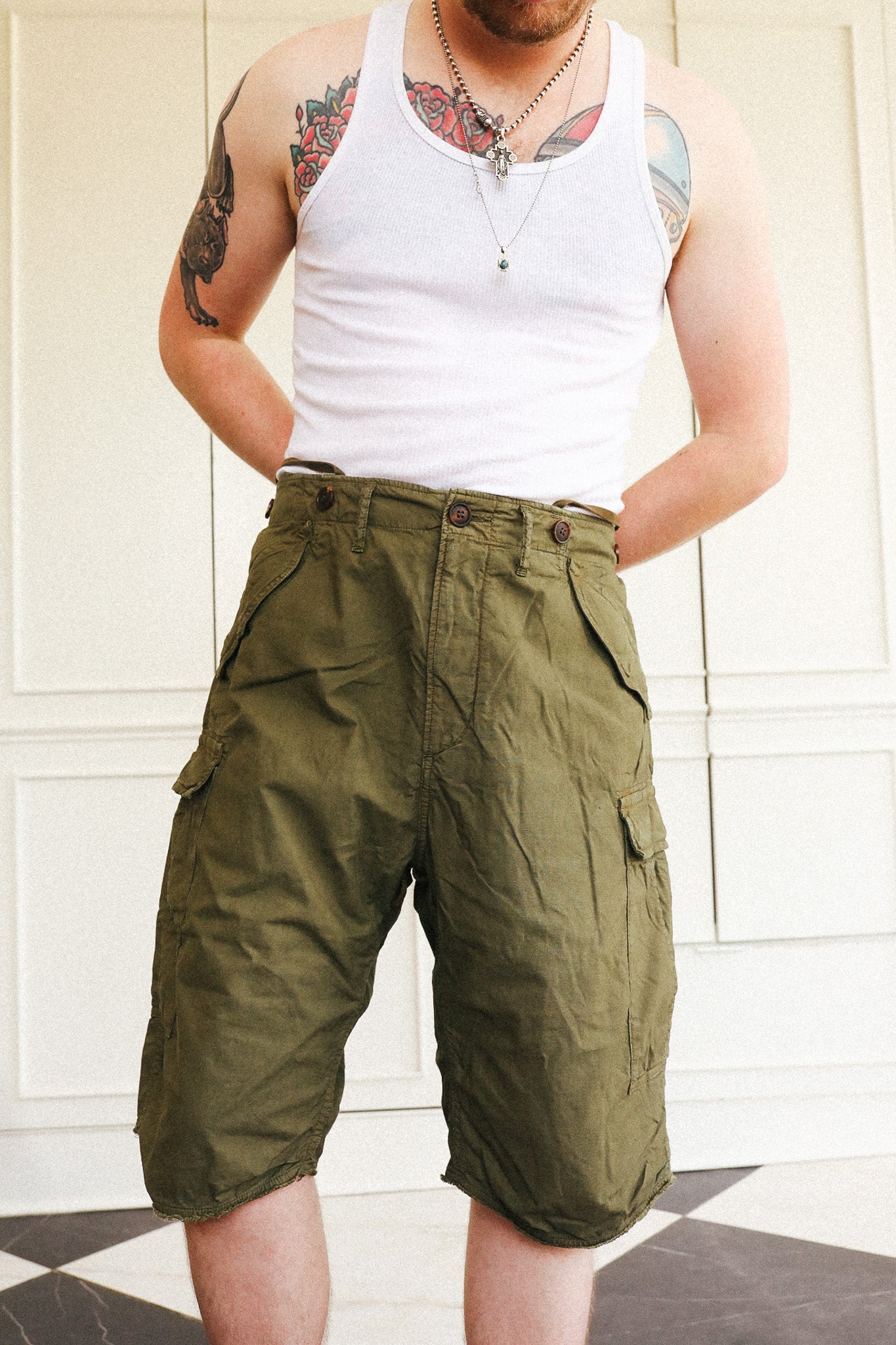 A man wearing olive green cargo shorts and a white tank top, standing in front of a white paneled wall.