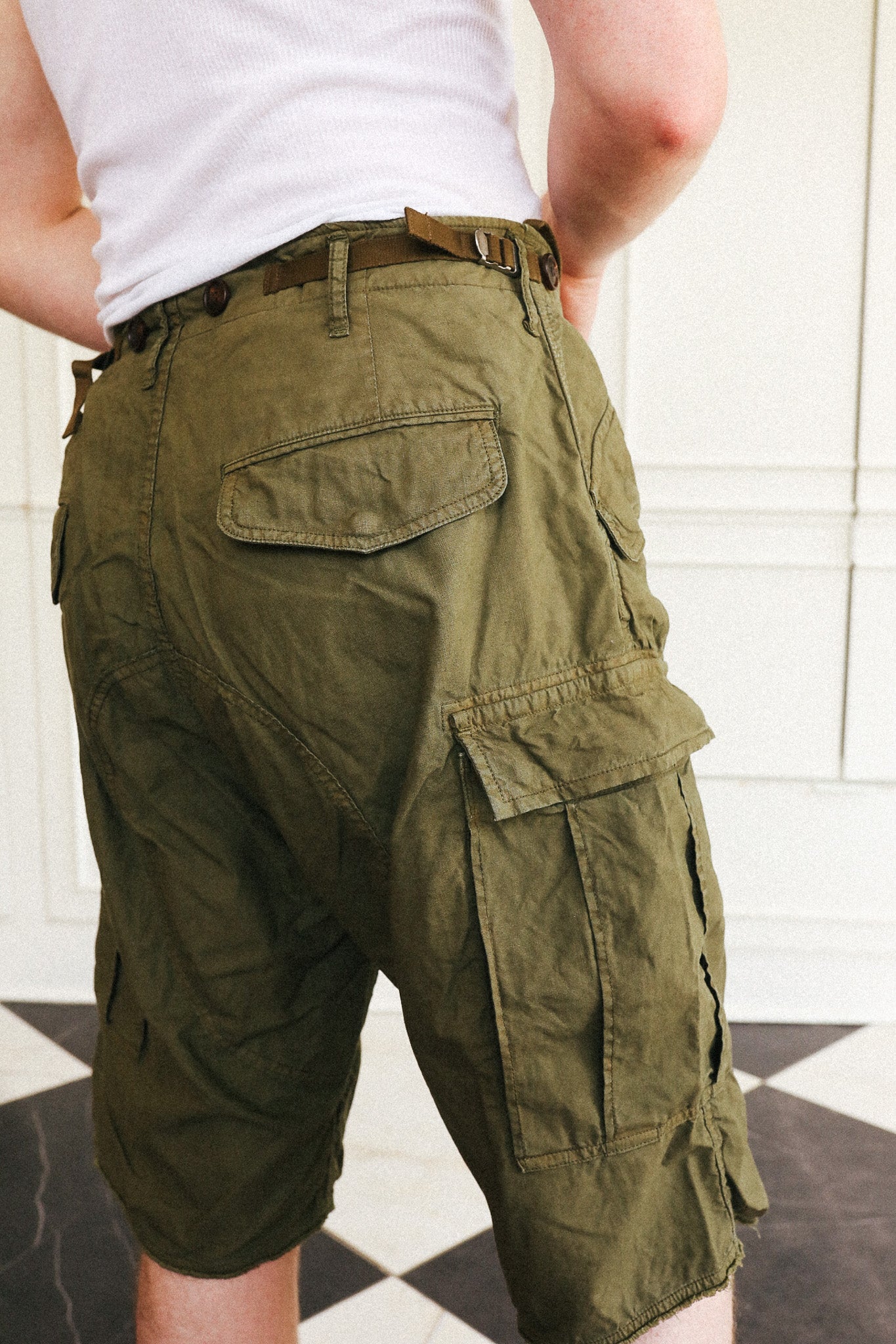 Olive green cargo shorts with a brown belt, shown from the back on a model wearing a white tank top, set against a white paneled wall and black-and-white checkered floor.
