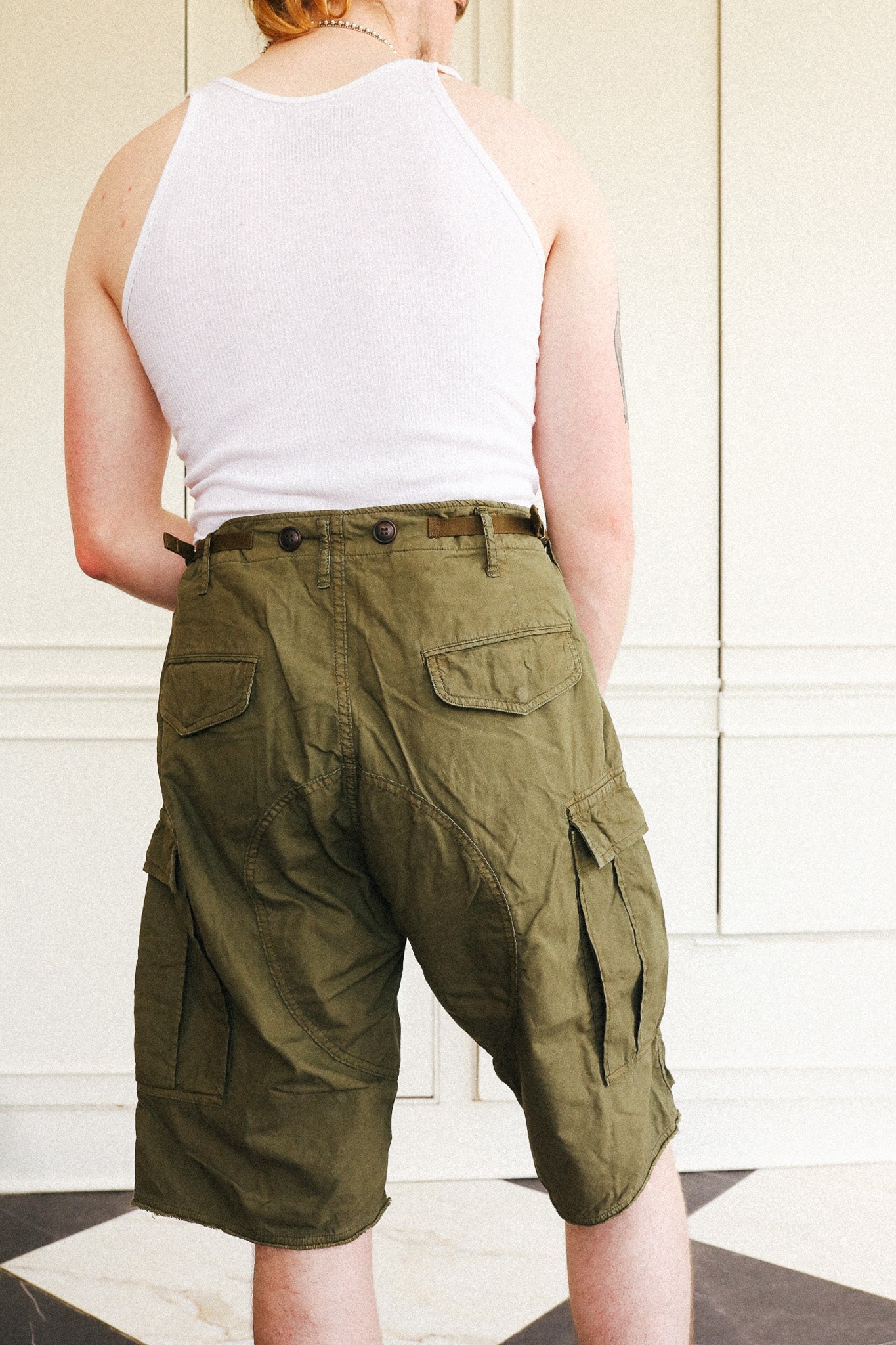 A person shown from the back wearing olive green cargo shorts and a white tank top, standing indoors against a light paneled wall.