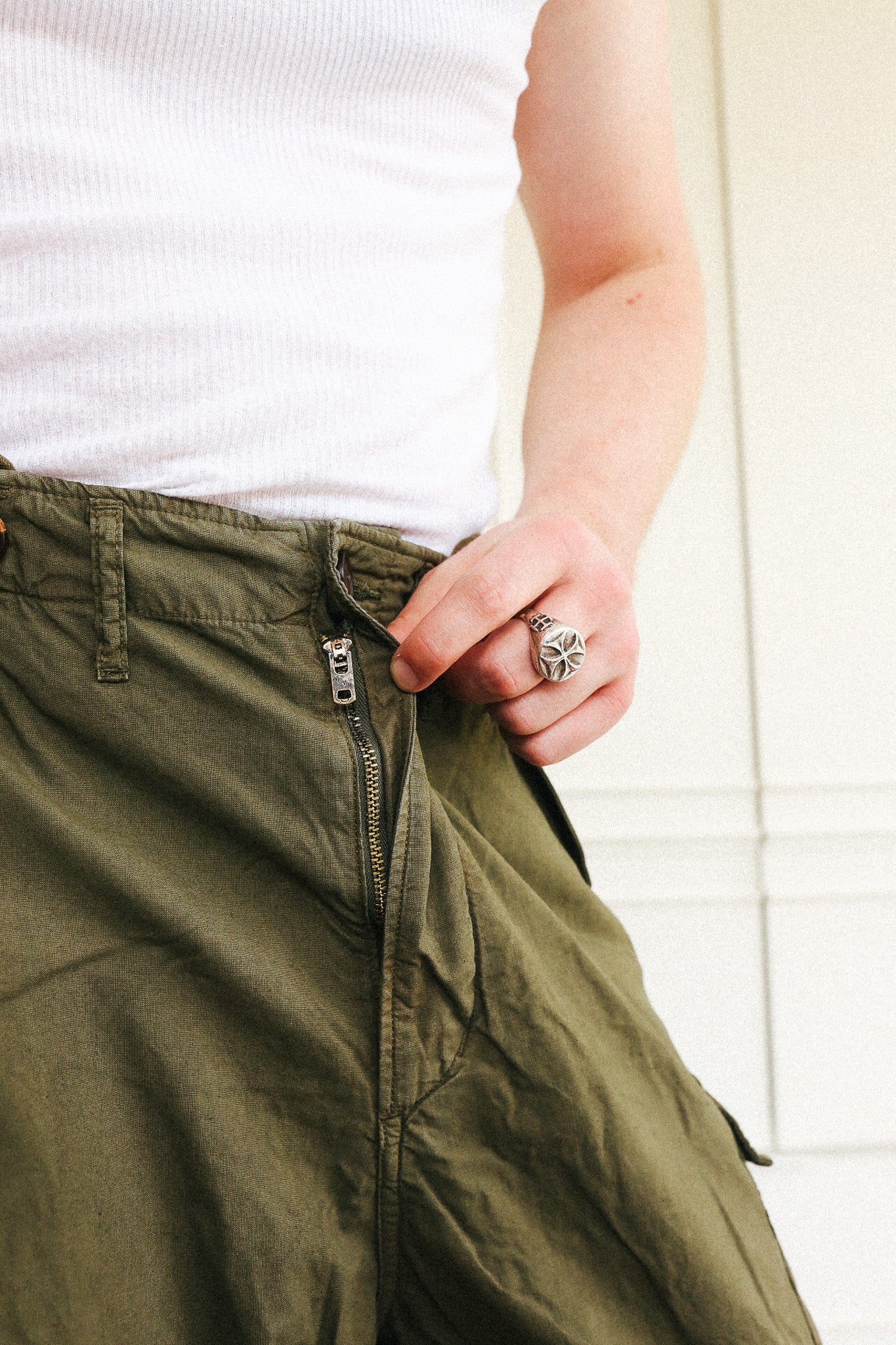 Close-up of a person wearing olive green cargo pants with a zipper fly and a white ribbed tank top, with a silver ring on their finger.