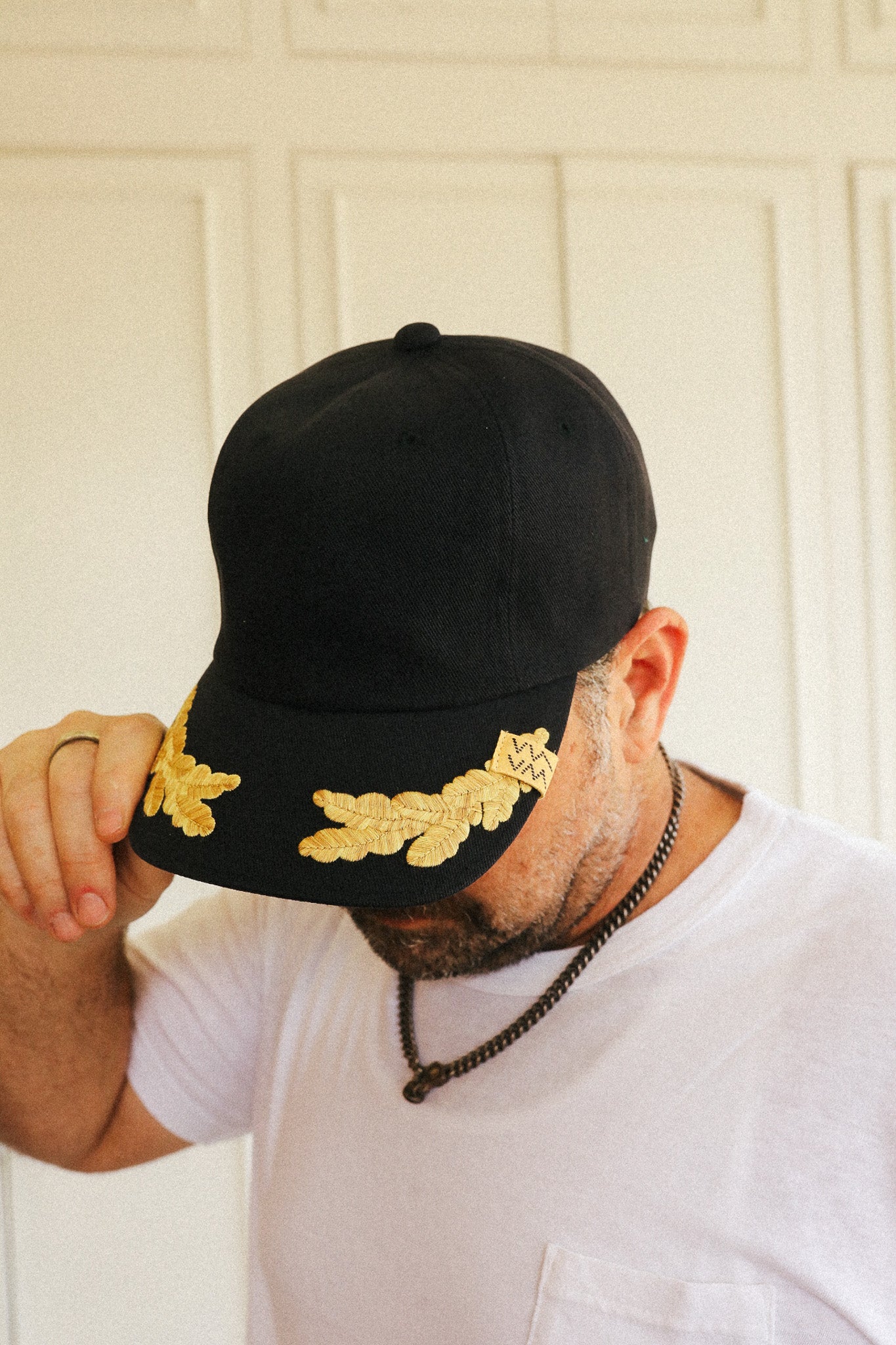 A man wearing a black baseball cap with gold embroidered laurel leaves on the brim, holding the brim down over his eyes. He is wearing a white t-shirt and a black beaded necklace, standing in front of a white paneled door.