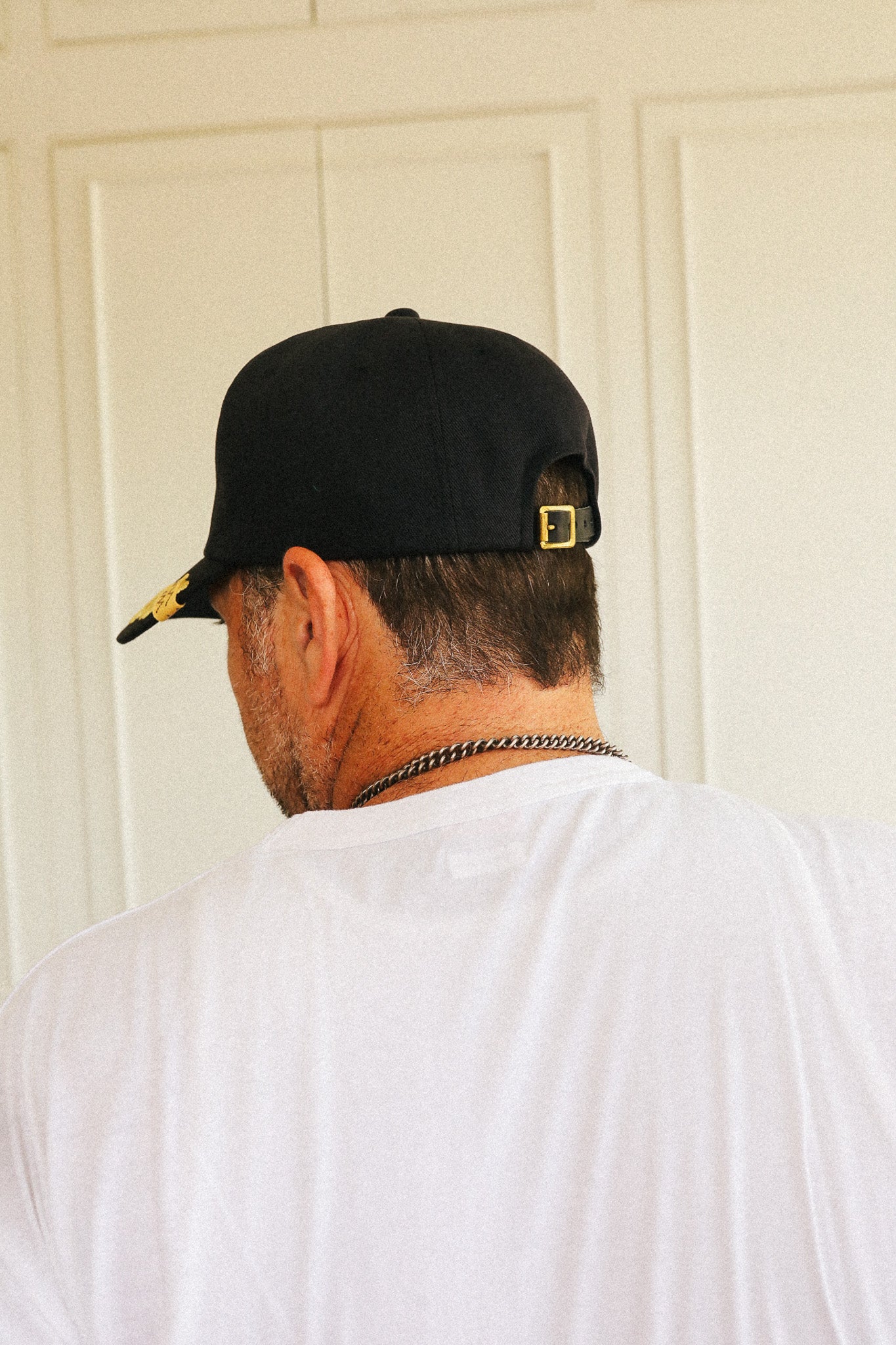 A man viewed from behind wearing a black baseball cap with a gold buckle and a white t-shirt, standing in front of a cream-colored paneled door.
