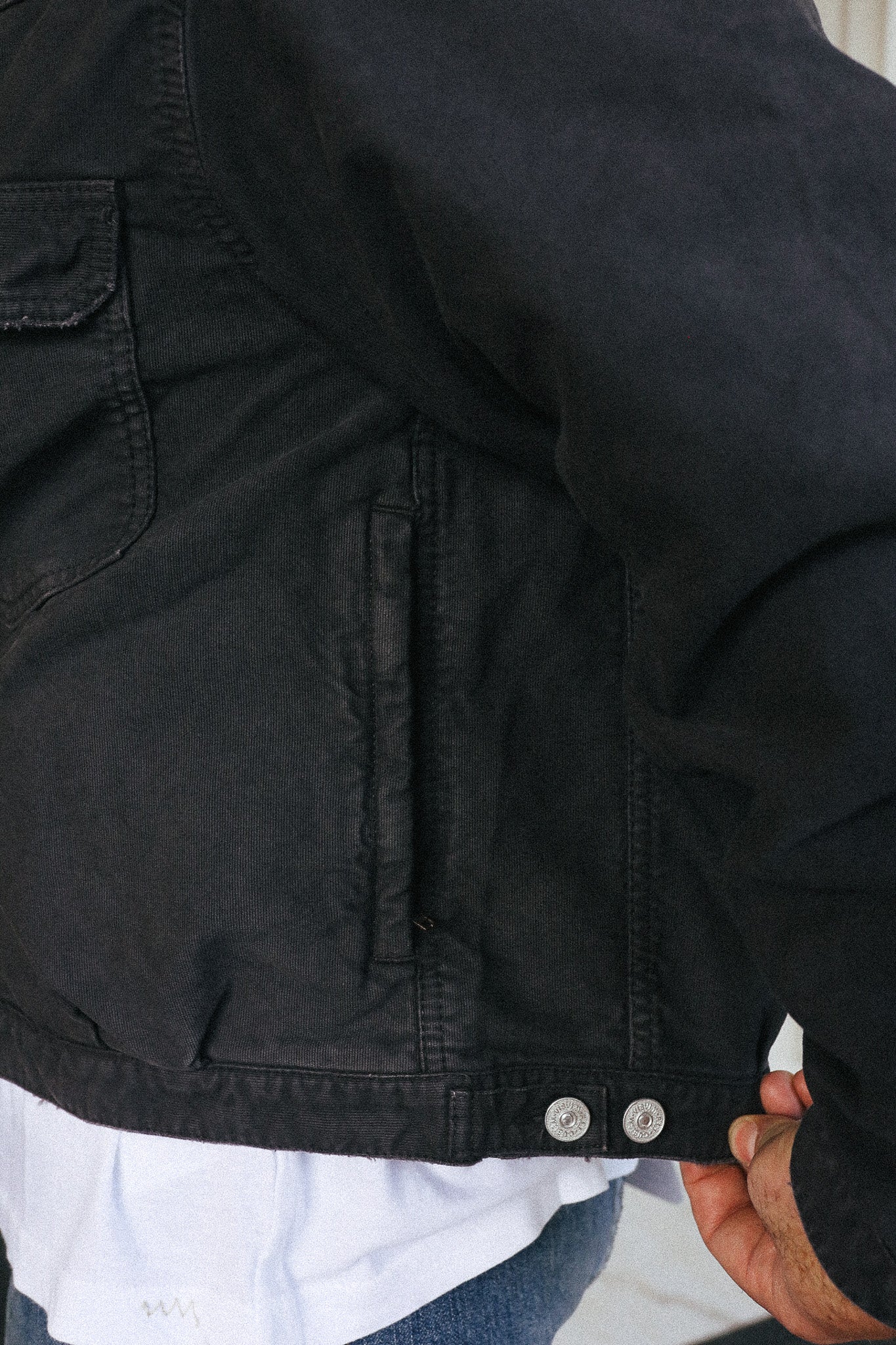 Close-up of a black canvas utility jacket with a hand adjusting the hem, worn over a white shirt.