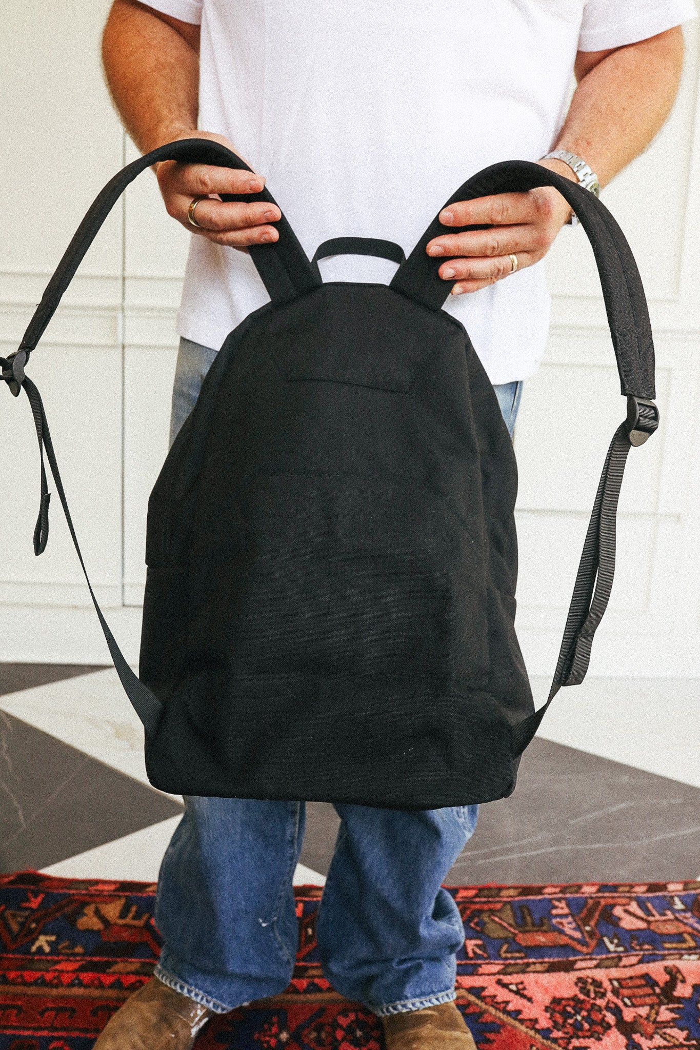 A person wearing a white t-shirt and blue jeans holds up a black canvas backpack against a white paneled wall.
