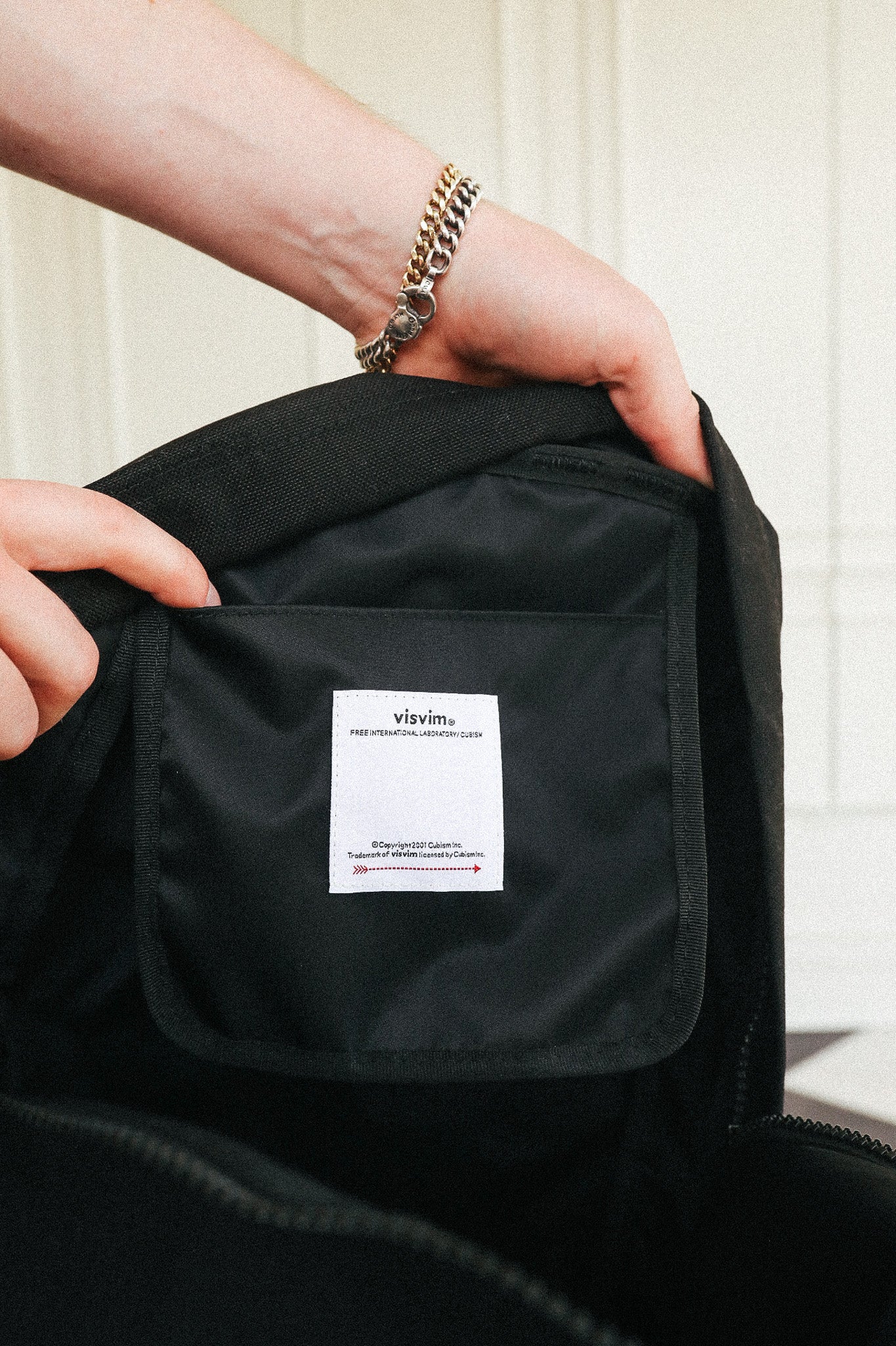 Close-up of a person's hands holding open a black backpack to show the interior pocket and a white sewn-in label that reads 'vayers'.