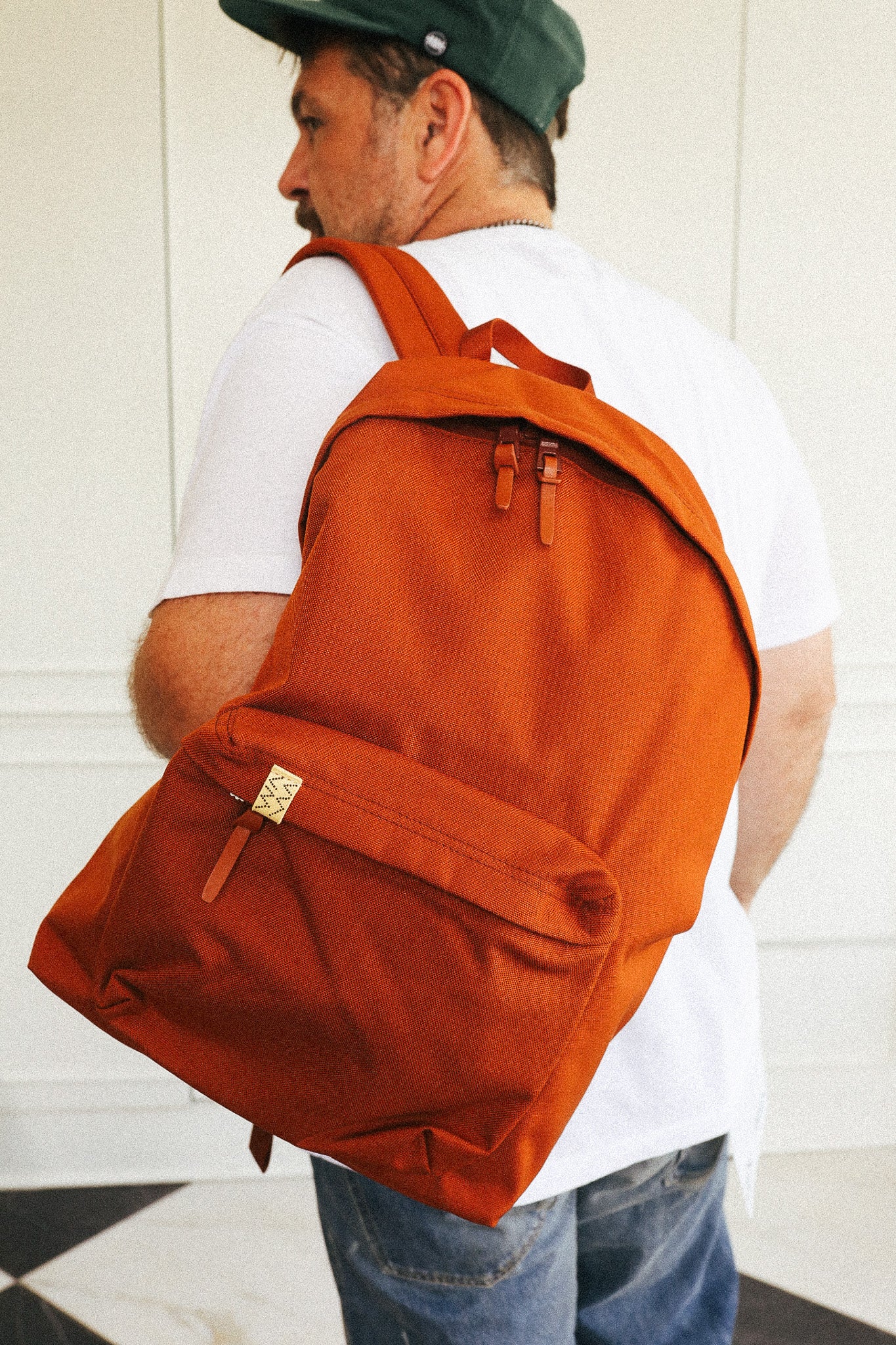 A man wearing a white t-shirt, blue jeans, and a green cap is shown from the back, carrying a large rust-orange canvas backpack with a front flap pocket and leather strap details.