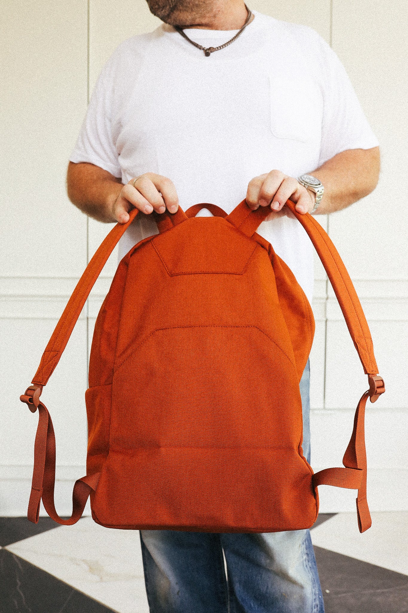 A man wearing a white t-shirt and blue jeans holds up an orange canvas backpack with brown straps in front of a white paneled wall.