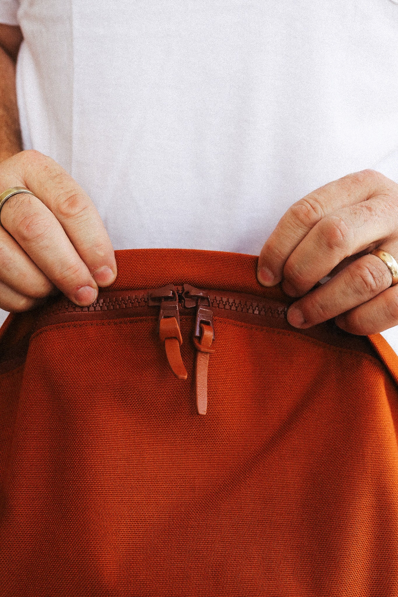 Close-up of a person's hands holding open a rust-orange canvas bag with a brown leather zipper pull, against a white shirt background.