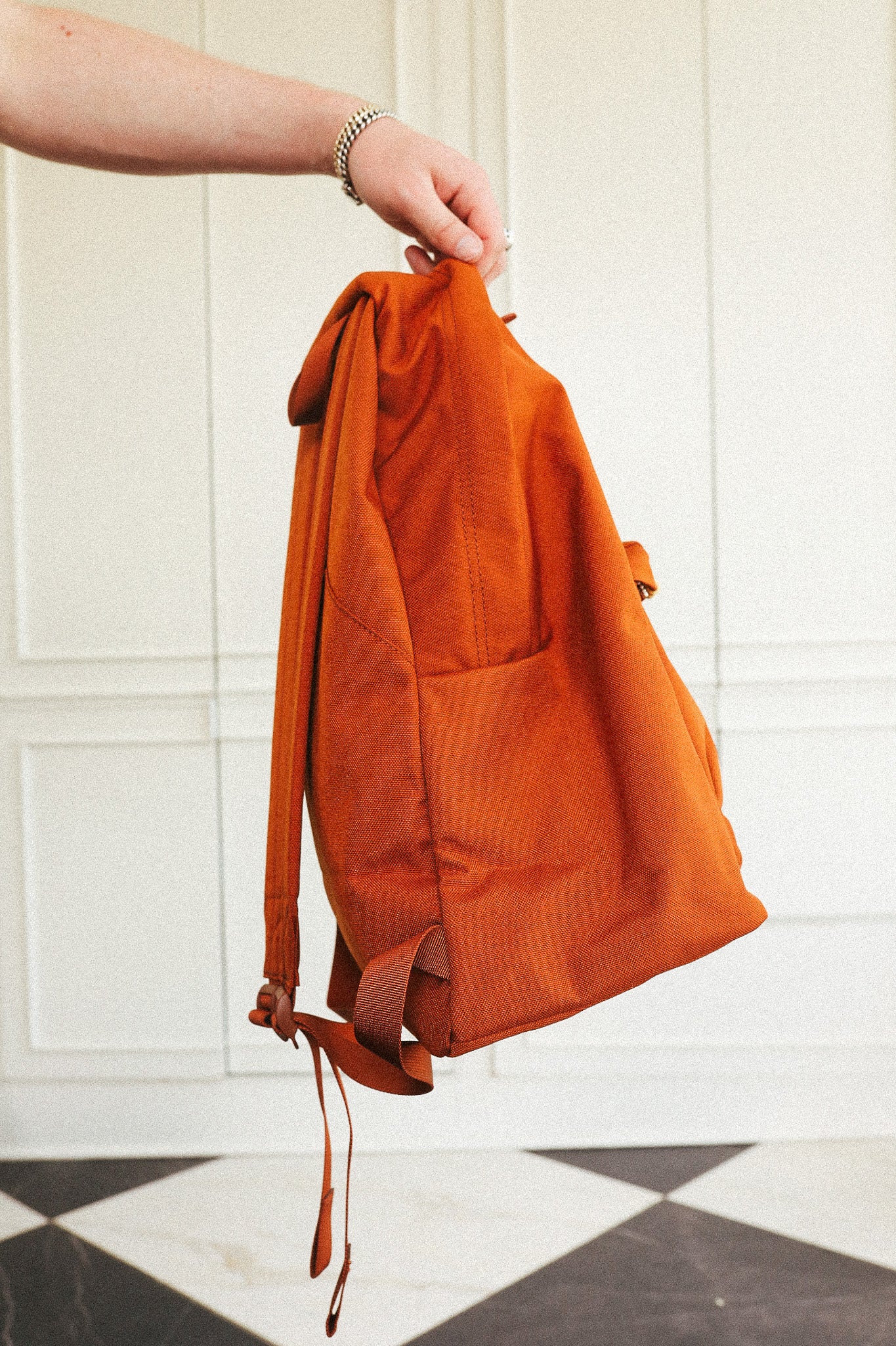 An orange canvas backpack held by a person's hand in front of a white paneled wall and black-and-white checkered floor.