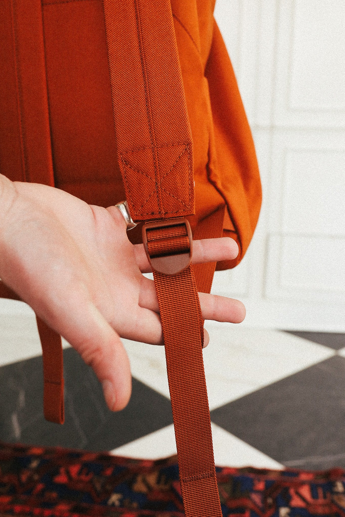 A close-up of a hand adjusting the strap buckle of an orange canvas backpack, with the backpack's fabric and a brown nylon strap visible.