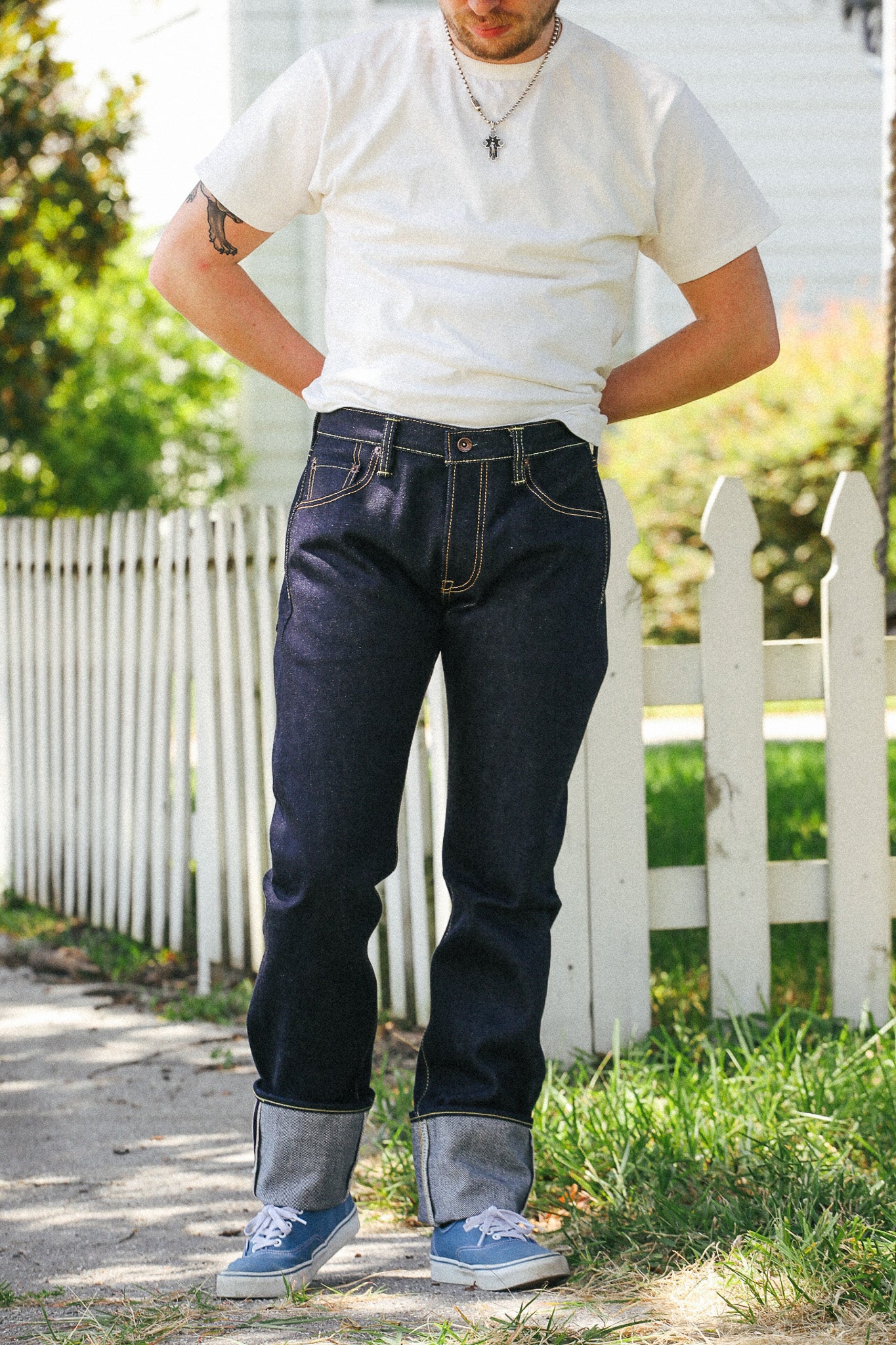 A man wearing dark wash blue jeans with cuffed hems, a white t-shirt, and blue sneakers, standing outdoors in front of a white picket fence.