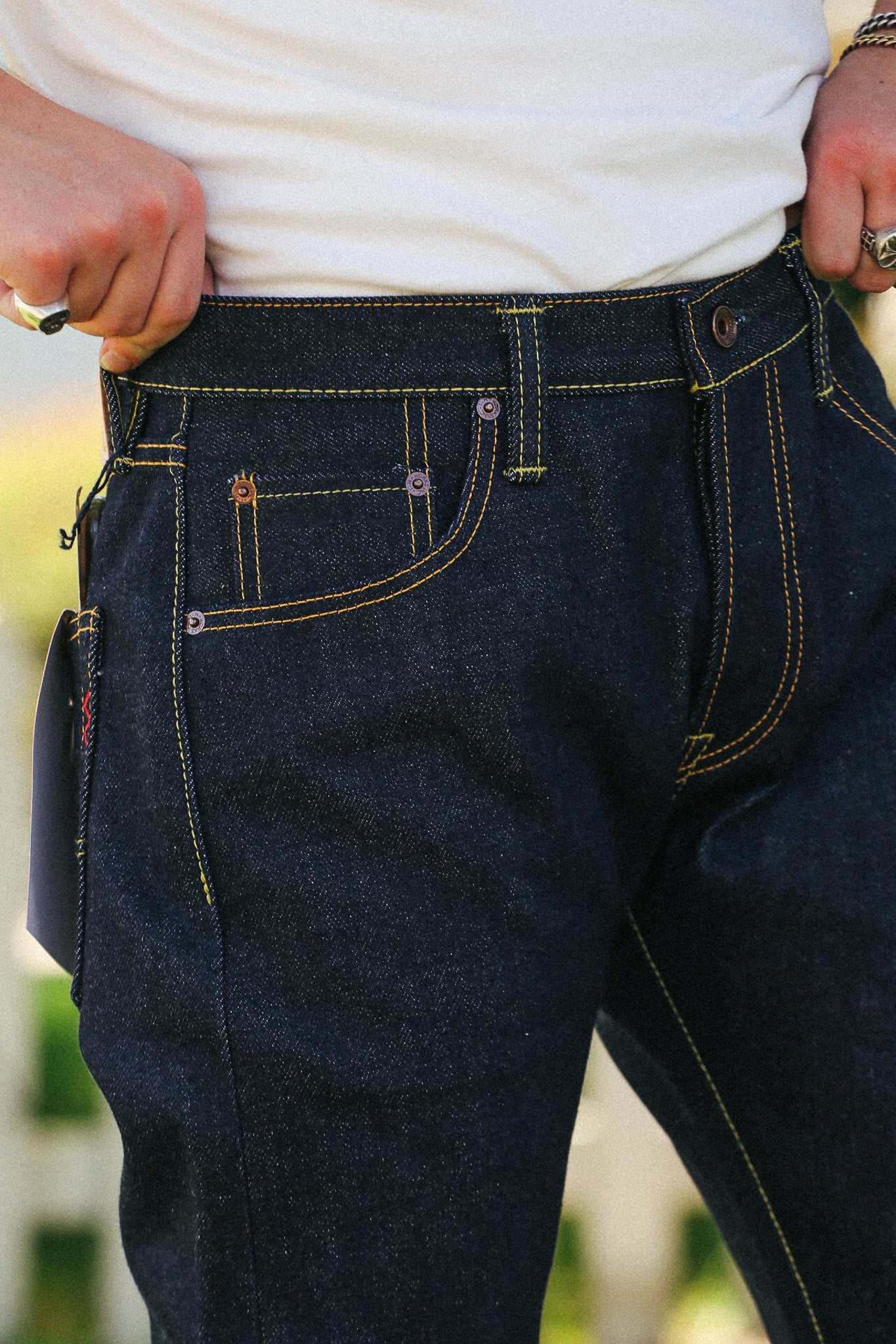 Close-up of a person wearing dark indigo denim jeans with contrasting yellow stitching, pulling the waistband with both hands. A white t-shirt is visible above the jeans.