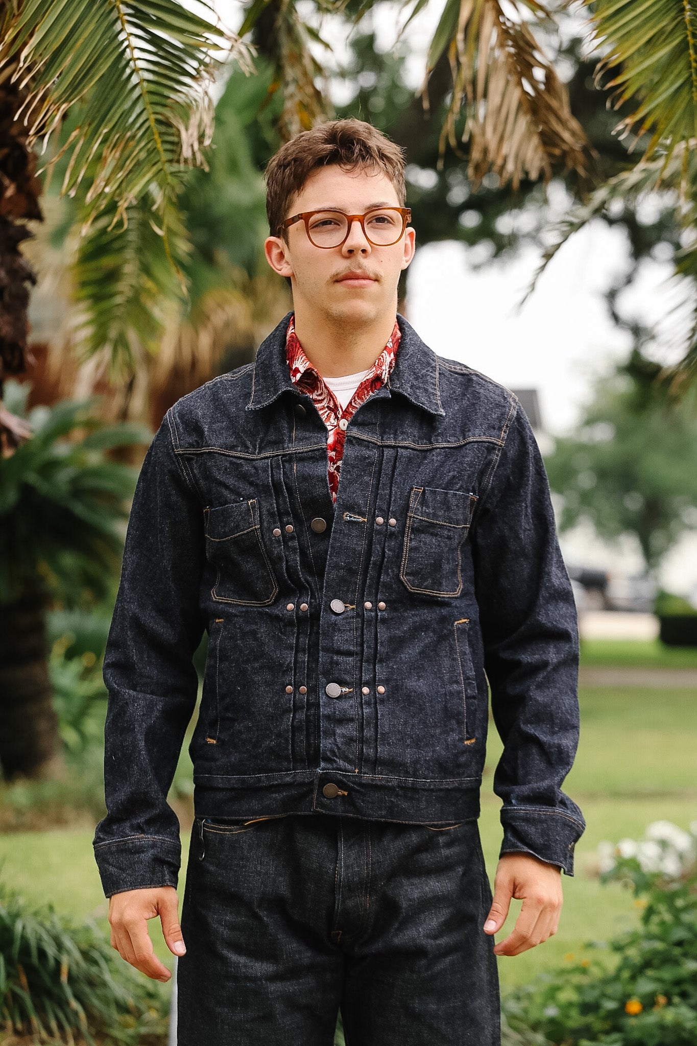 Person wearing a denim jacket and glasses standing outdoors with greenery in the background