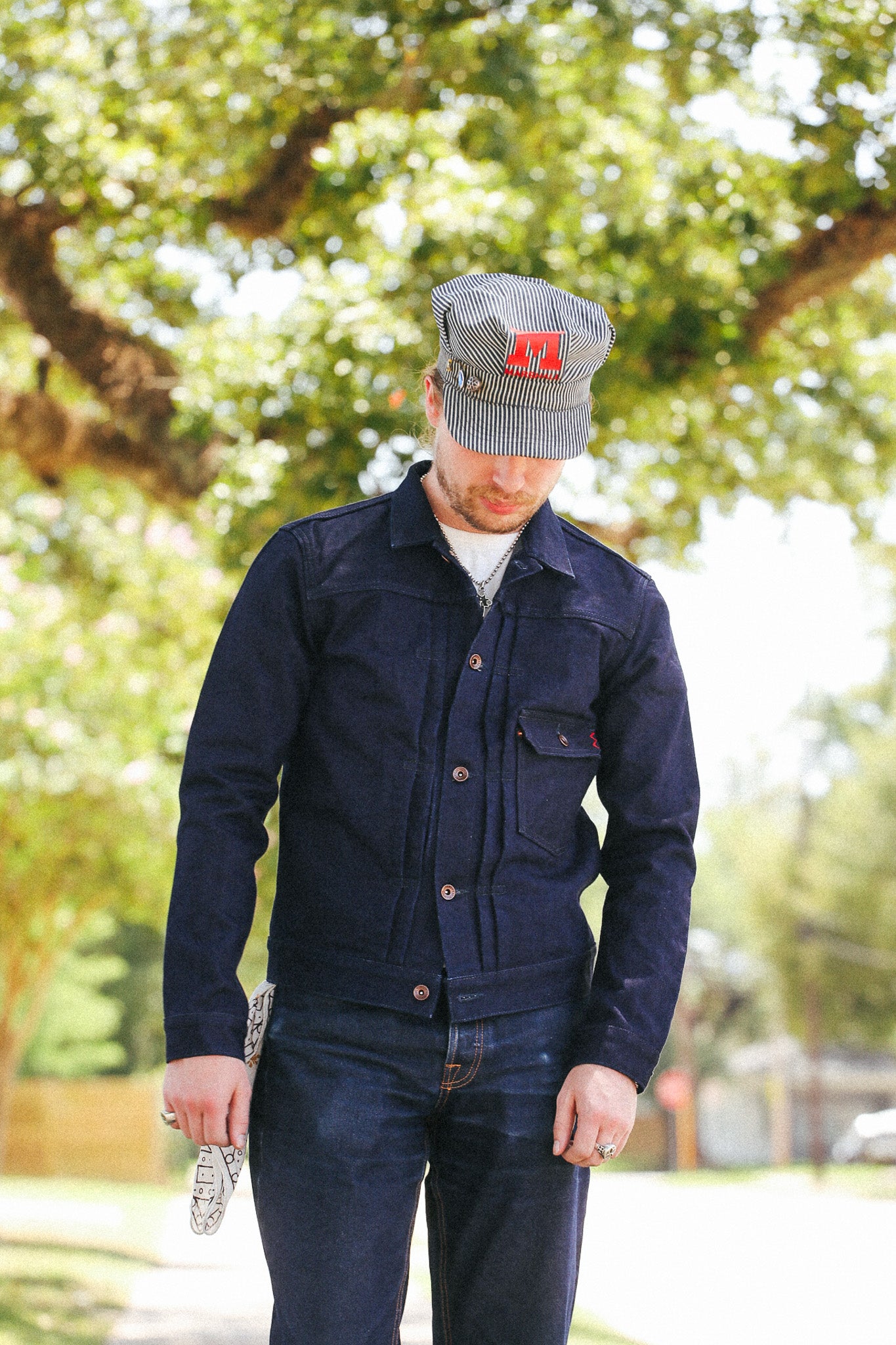 A man wearing a dark navy blue corduroy jacket and matching dark jeans, standing outdoors in front of a blurred background of green trees. He is wearing a gray corduroy cap with a red embroidered logo.