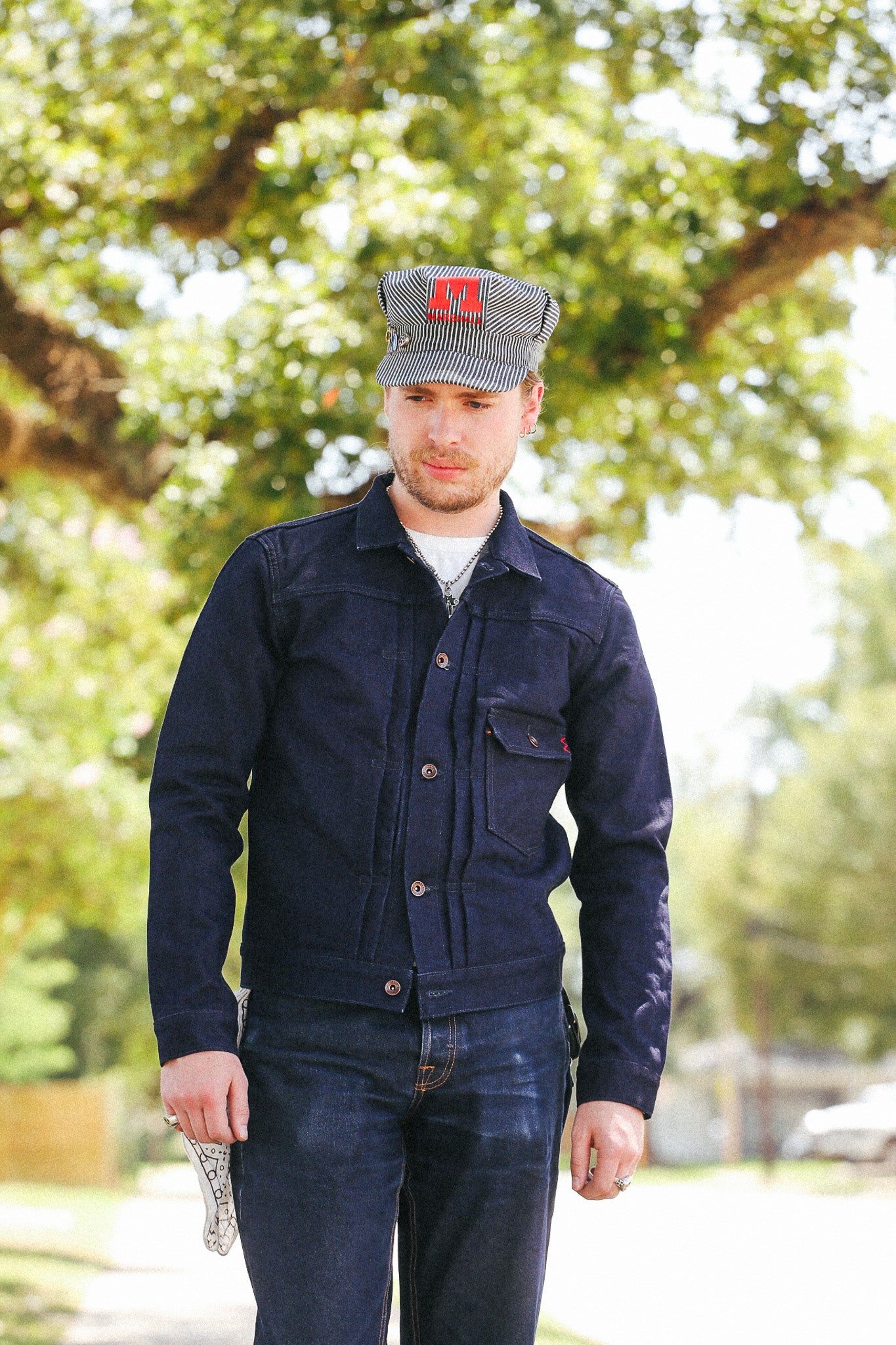 A man wearing a dark navy blue denim work jacket, matching dark jeans, and a gray cap with a red logo, standing outdoors in front of a blurred green tree background.