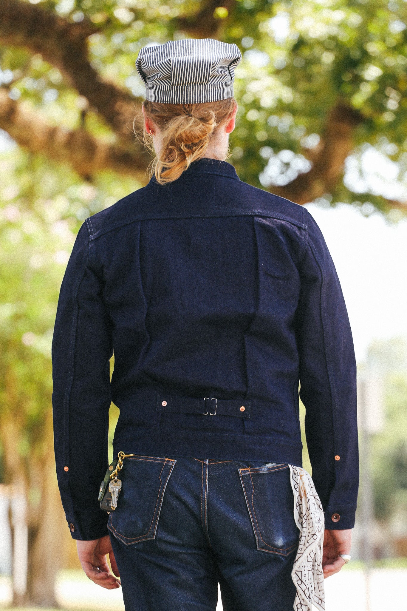 A person viewed from behind wearing a dark indigo denim work jacket with a matching striped cap, standing outdoors in front of a blurred background of trees.