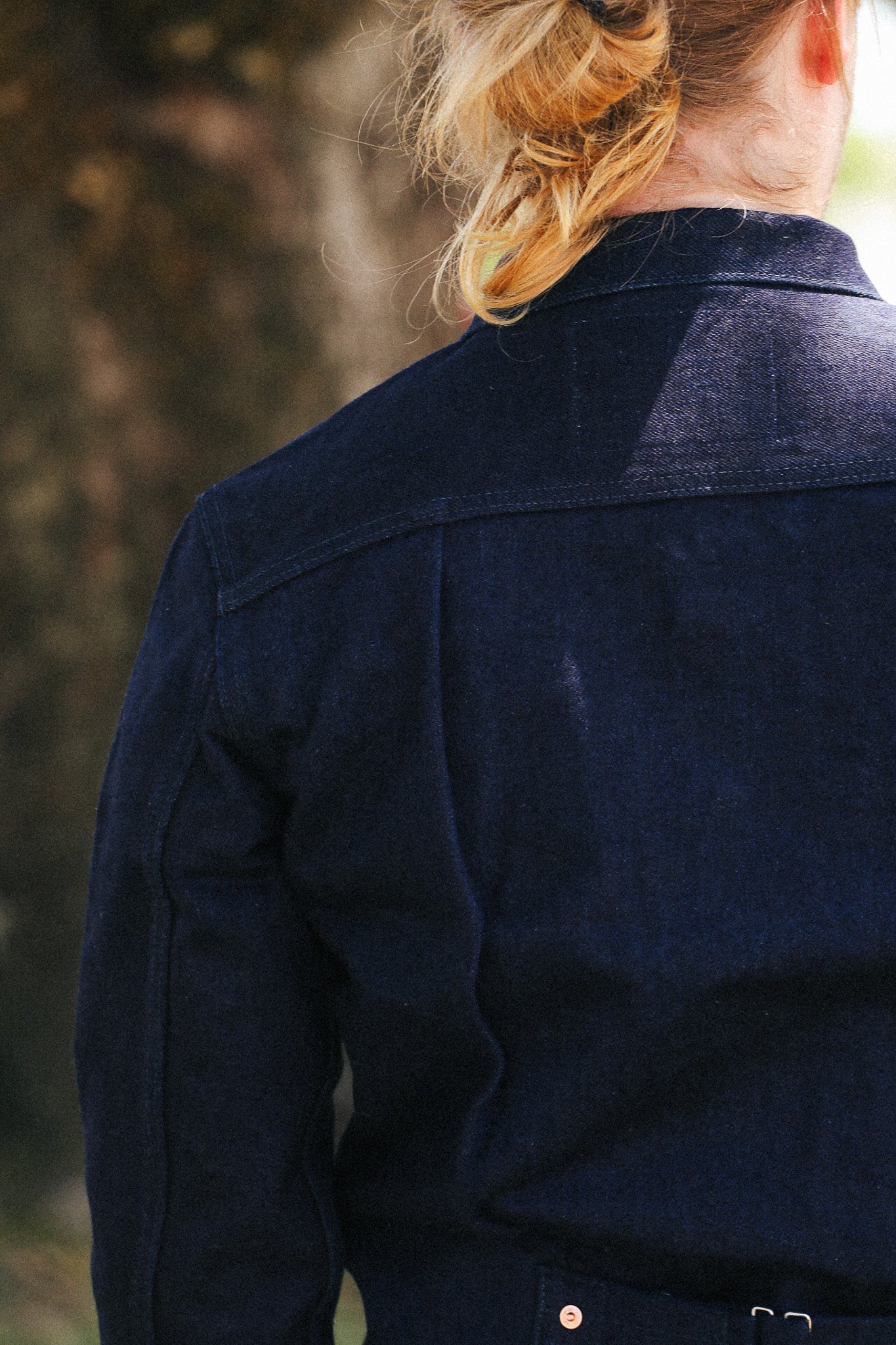 Back view of a person wearing a dark indigo denim jacket outdoors, with the hair tied back in a bun.