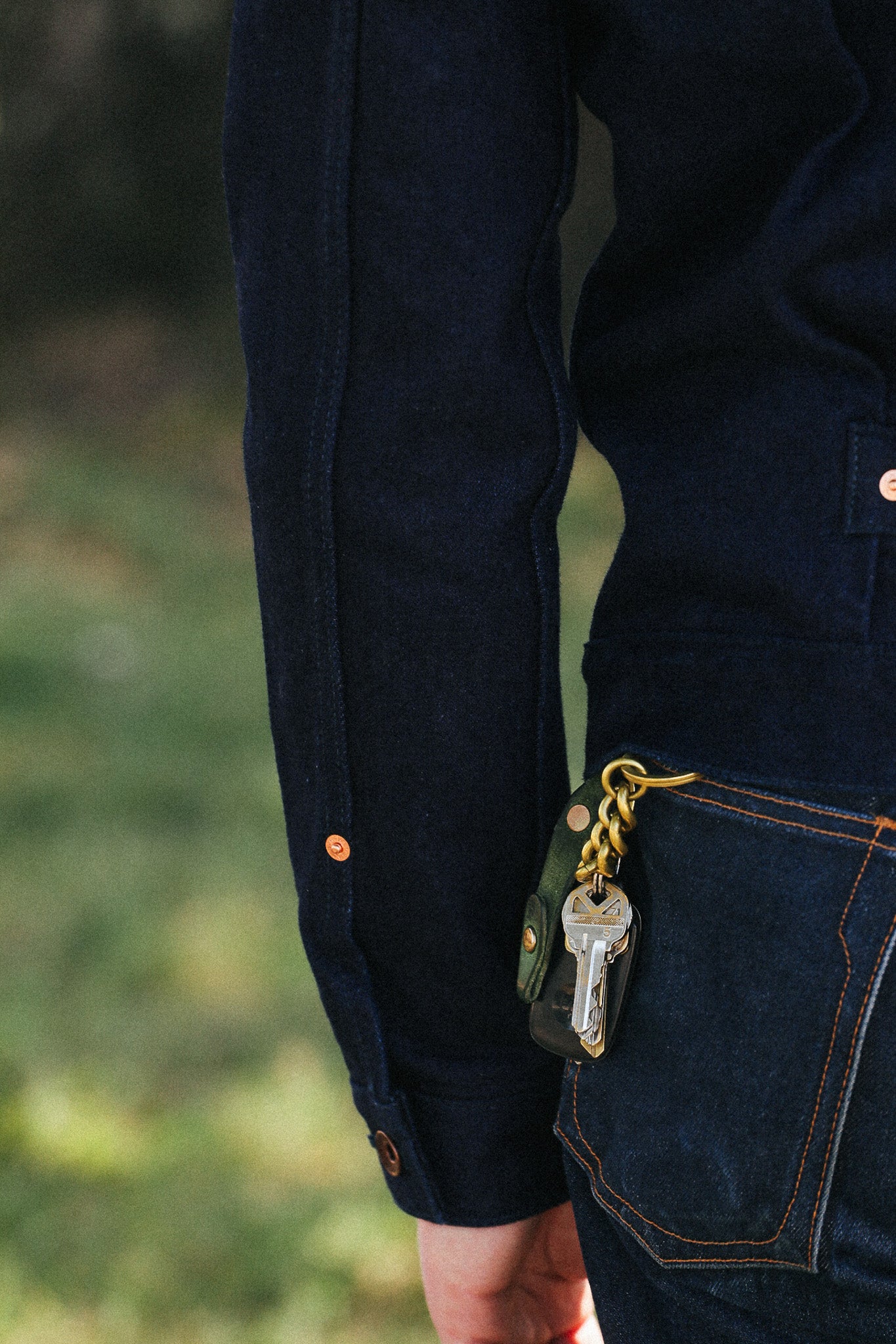 Close-up of a person wearing dark indigo denim overalls, with a key fob attached to the back pocket, against a blurred outdoor background.