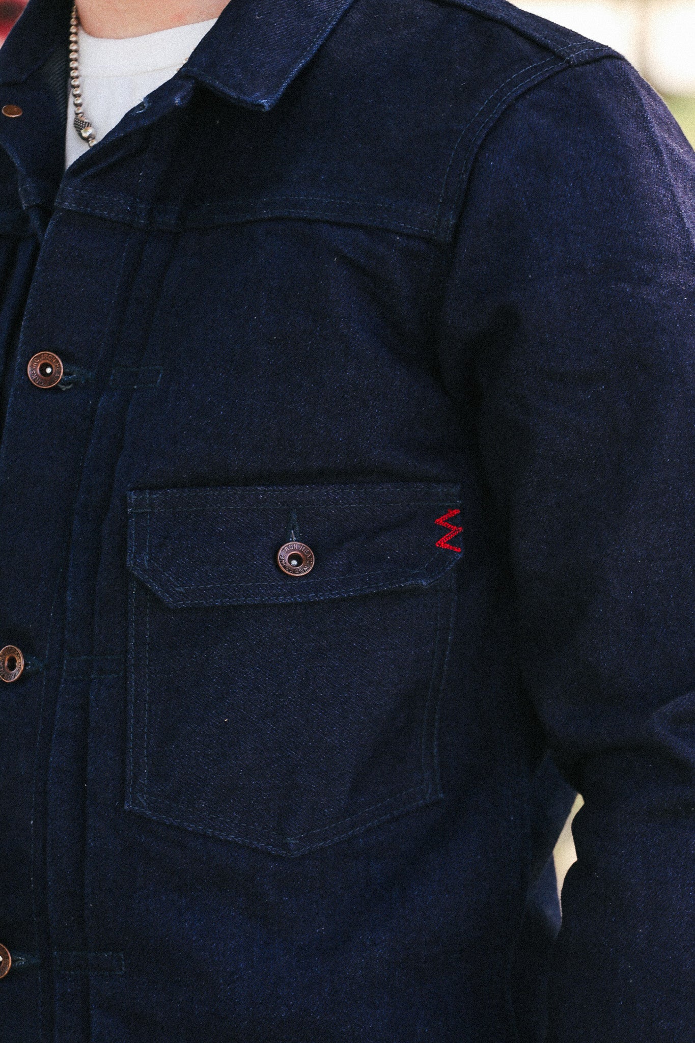 Close-up of a person wearing a dark indigo denim jacket with a chest pocket, brown buttons, and a small red zigzag embroidery detail on the pocket flap.