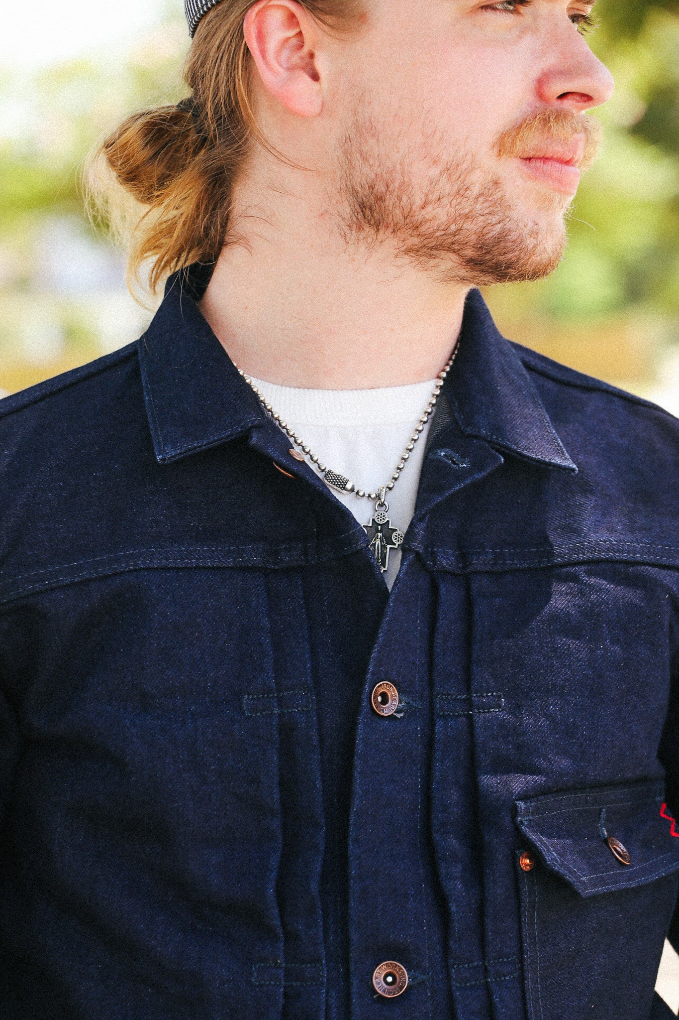 A close-up side profile of a man wearing a dark indigo denim work shirt over a white t-shirt, with a silver chain necklace and a dark beanie.
