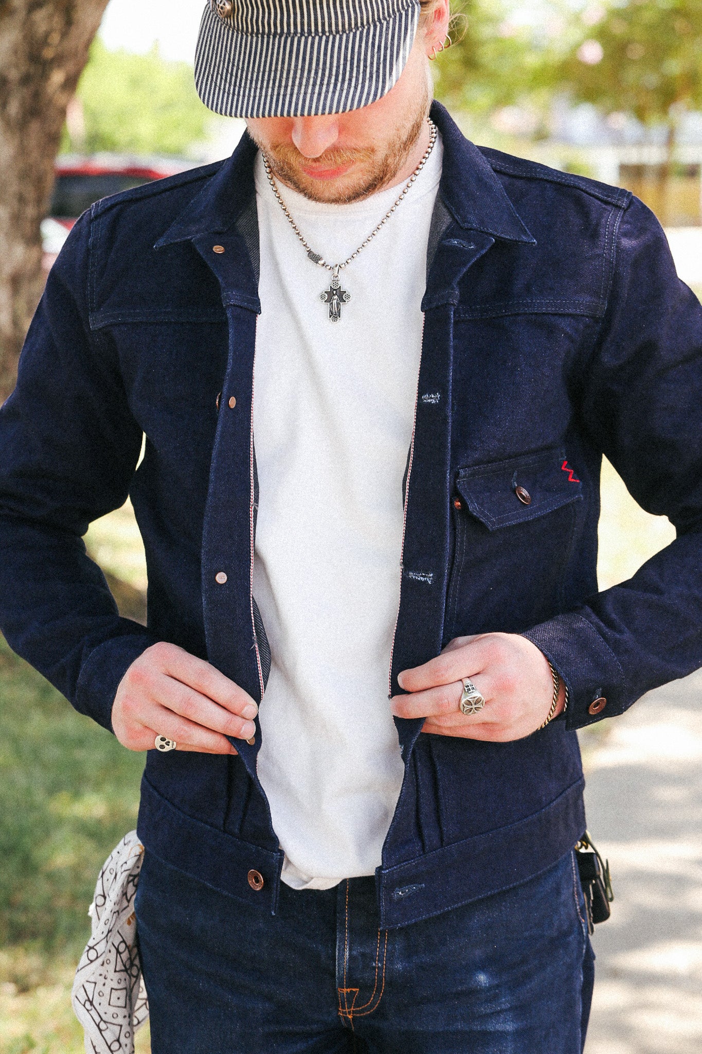 A man wearing a dark navy blue denim work jacket over a white t-shirt, paired with a striped cap and jeans, standing outdoors.