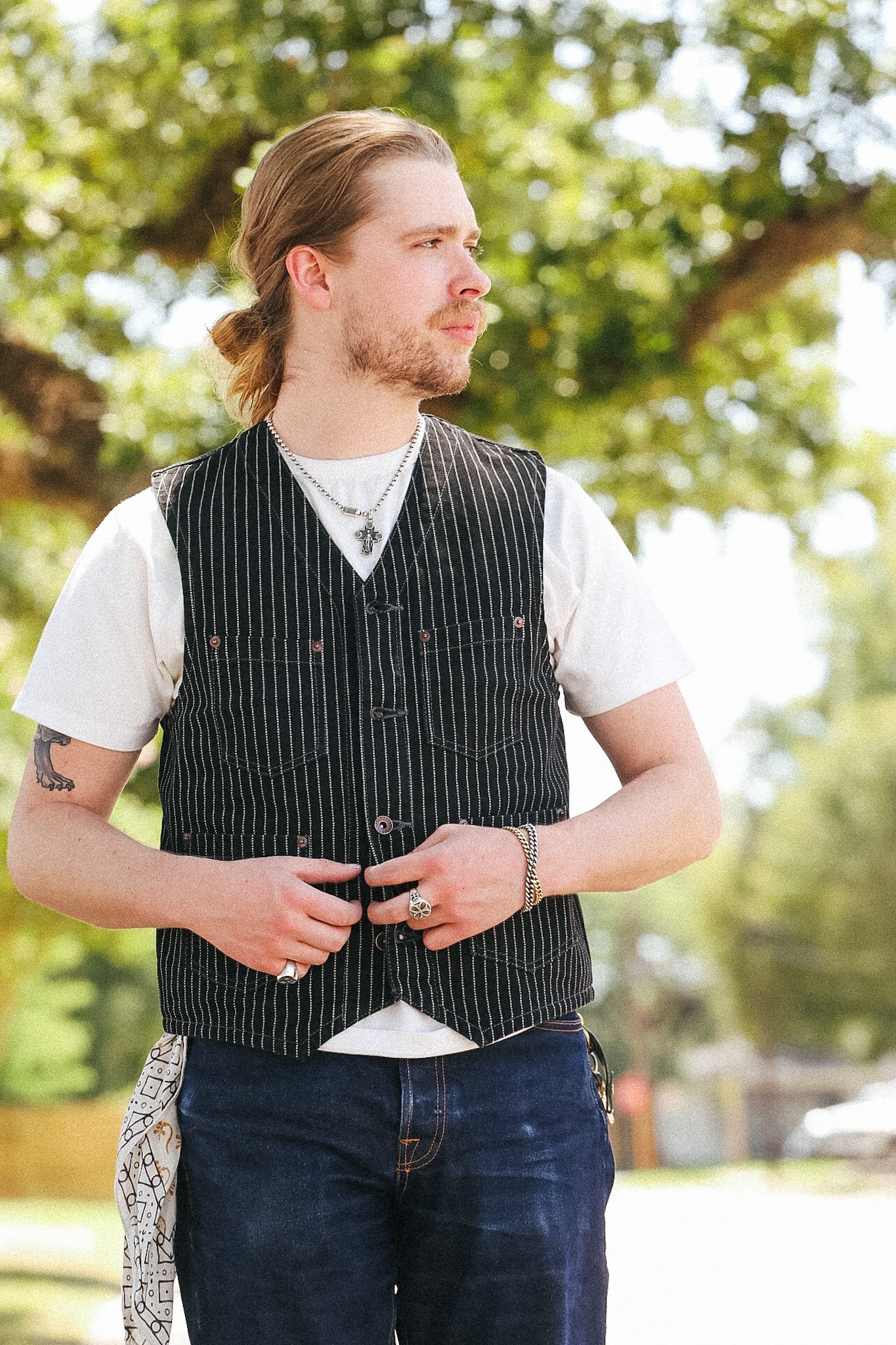 A man wearing a black pinstriped vest over a white t-shirt and dark blue jeans, standing outdoors in front of a blurred green tree background.