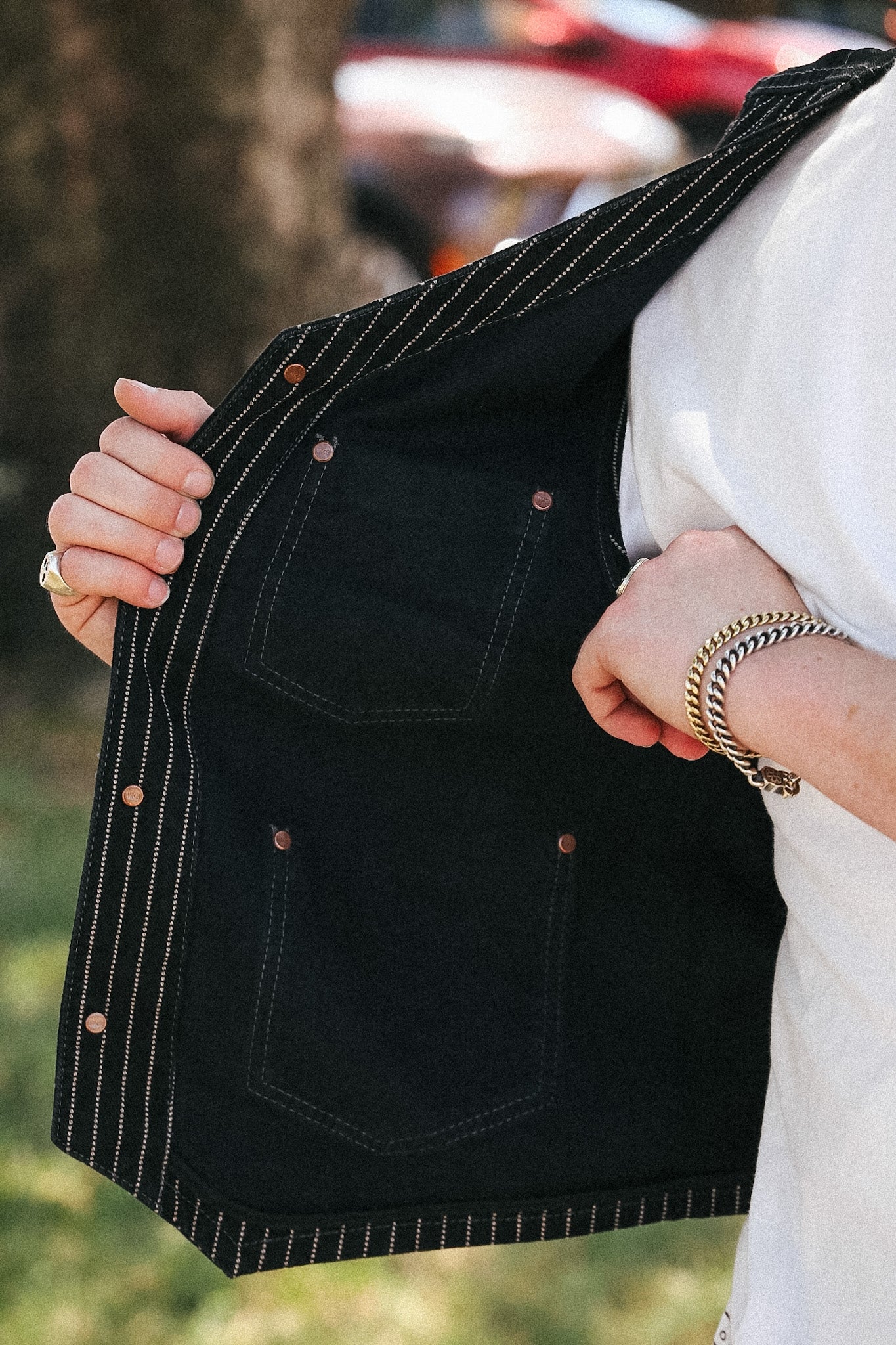 A close-up of a person wearing a black denim vest with thin gold pinstripes and copper-toned rivets, holding the vest open to show the interior pockets and lining. The person is also wearing a white shirt and gold bracelets, with a blurred outdoor background.