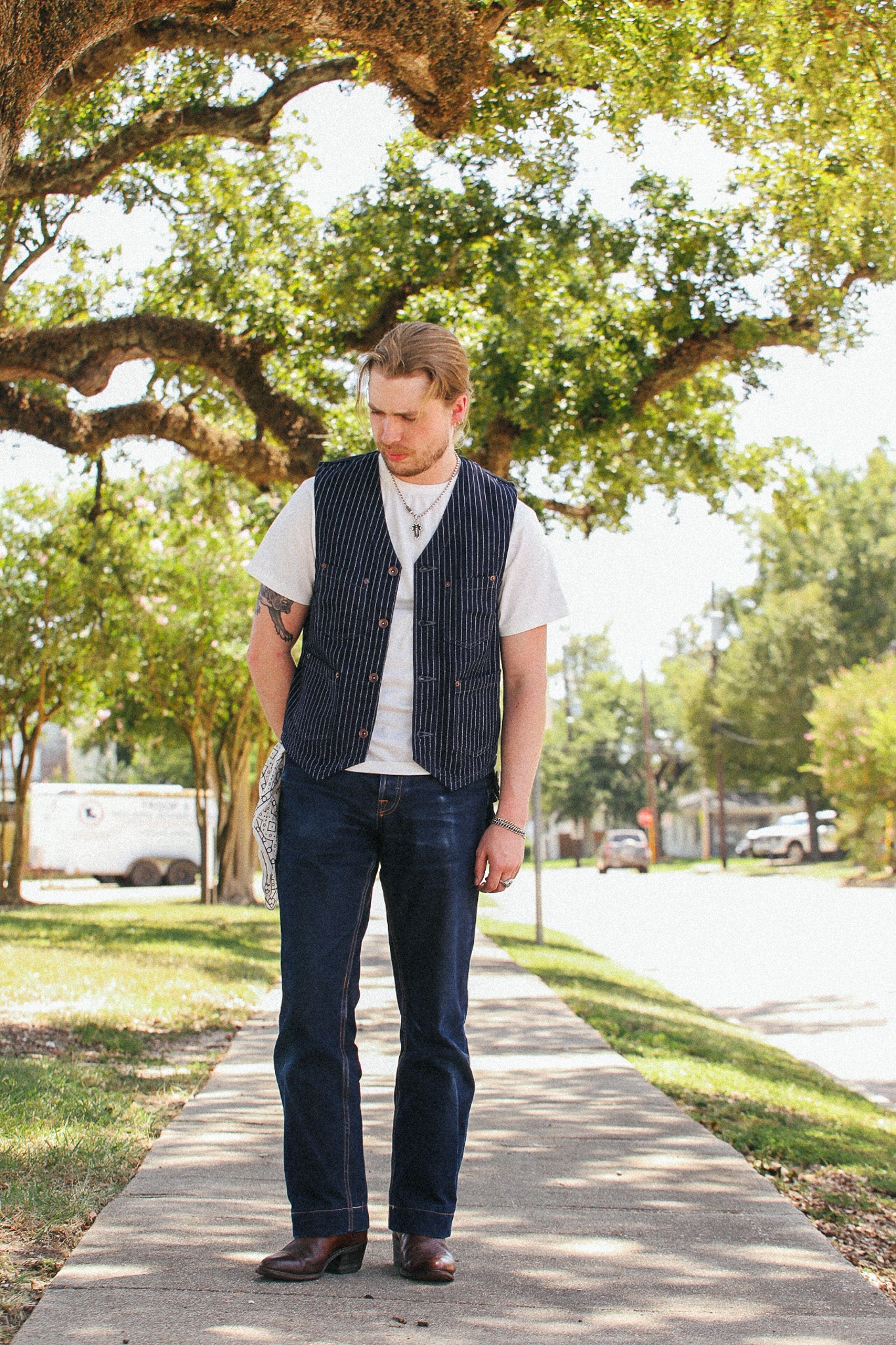 A man standing on a sidewalk outdoors under a large tree, wearing a white t-shirt, a dark navy pinstriped vest, dark blue jeans, and brown boots.