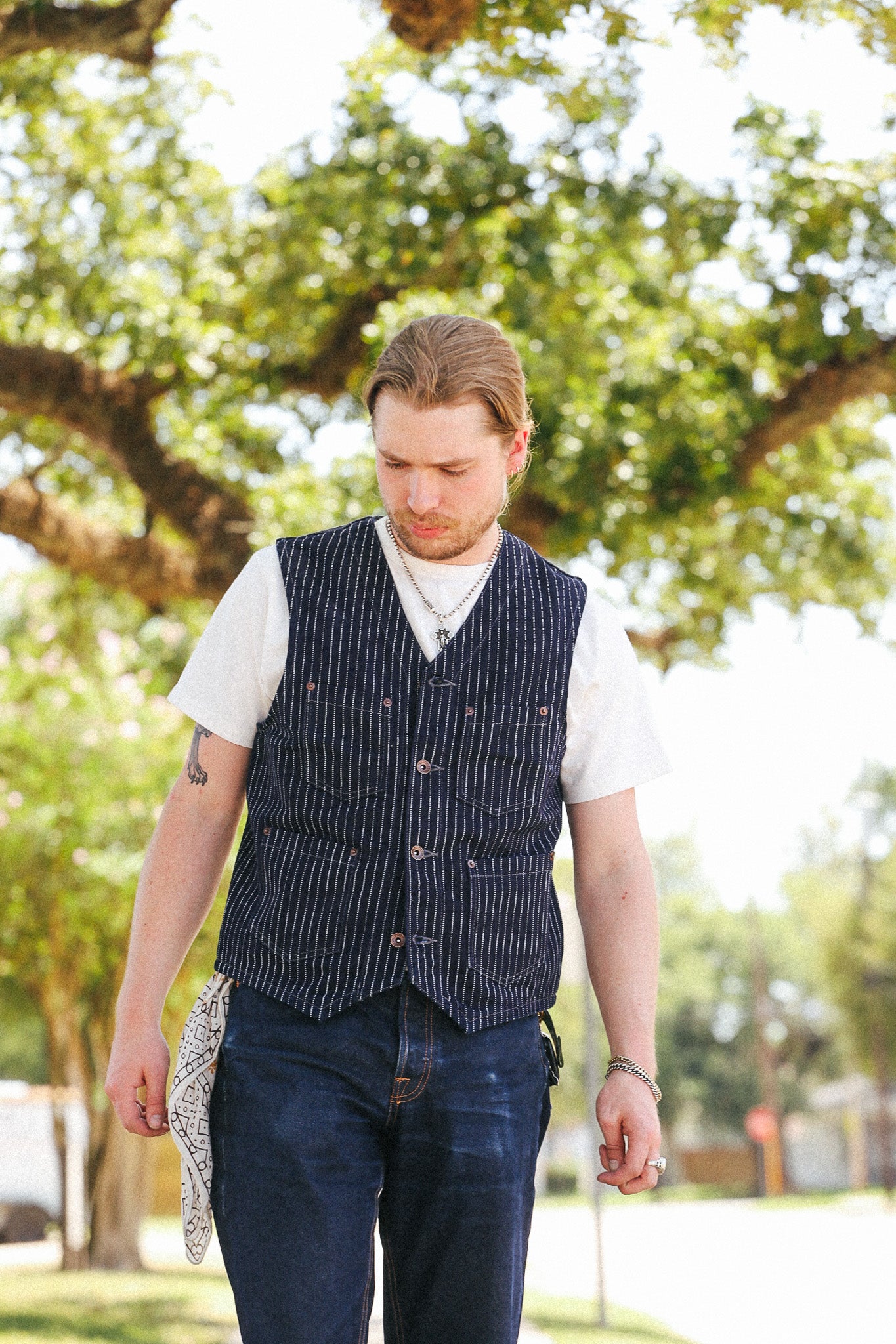 A man walking outdoors in a park wearing a navy blue pinstriped vest over a white t-shirt and dark blue jeans, holding a patterned hat in one hand.