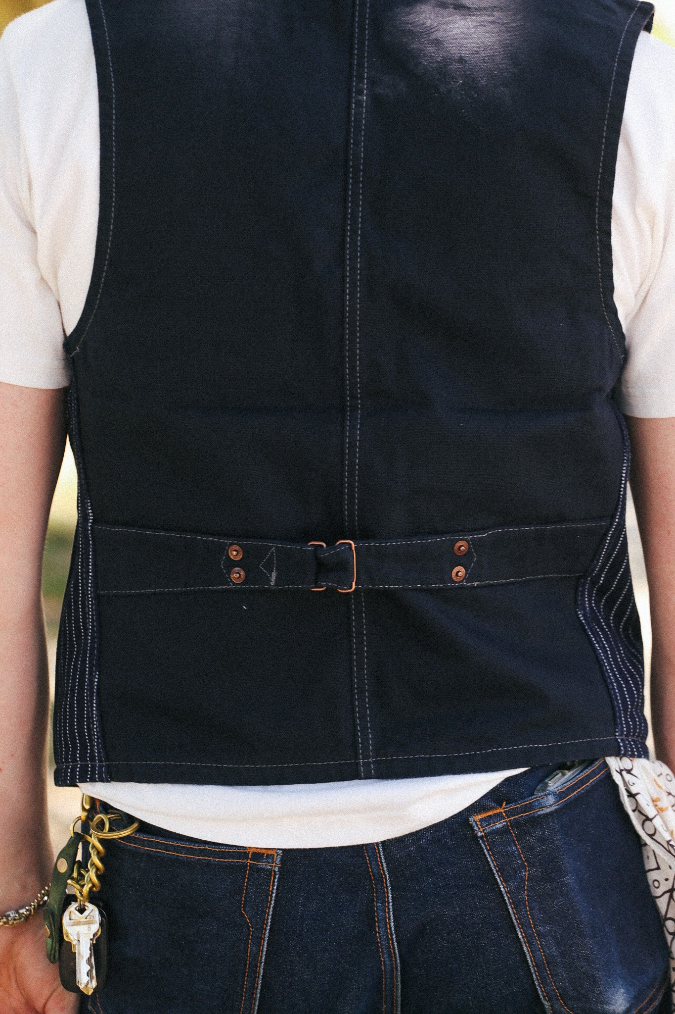 Close-up back view of a person wearing a dark indigo denim vest over a white t-shirt and dark blue jeans, with a belt and buckle detail at the waist.