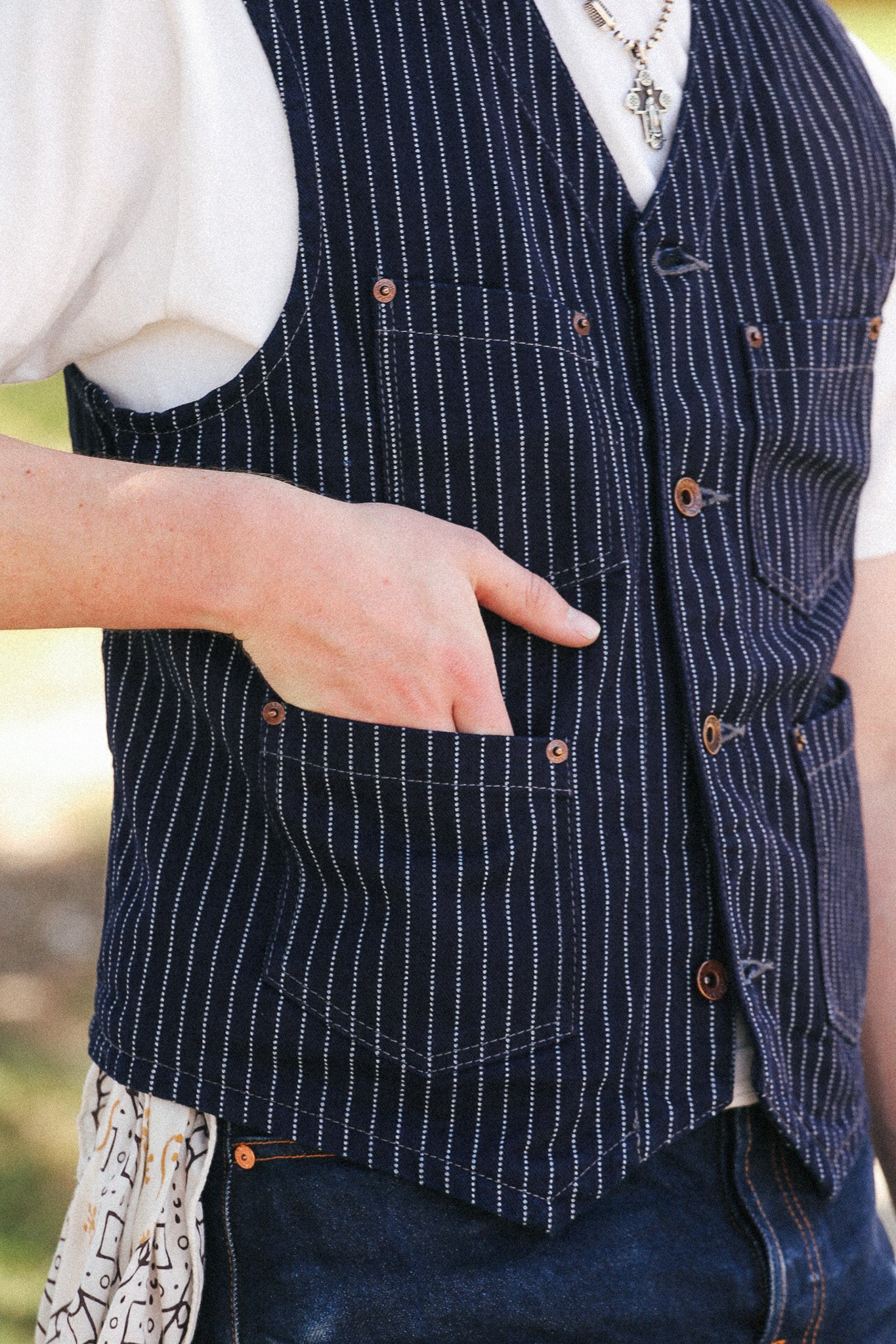 A close-up of a person wearing a navy blue vest with thin white vertical pinstripes, brown buttons, and front pockets. The person is also wearing a white shirt, dark blue jeans, and a patterned undershirt peeking out at the sides.