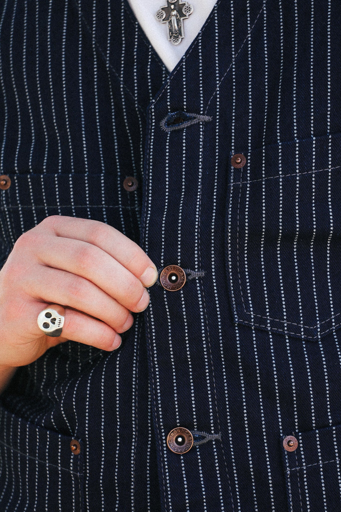 Close-up of a person wearing a dark navy pinstripe vest over a white collared shirt, with a silver cross necklace and a skull ring visible.